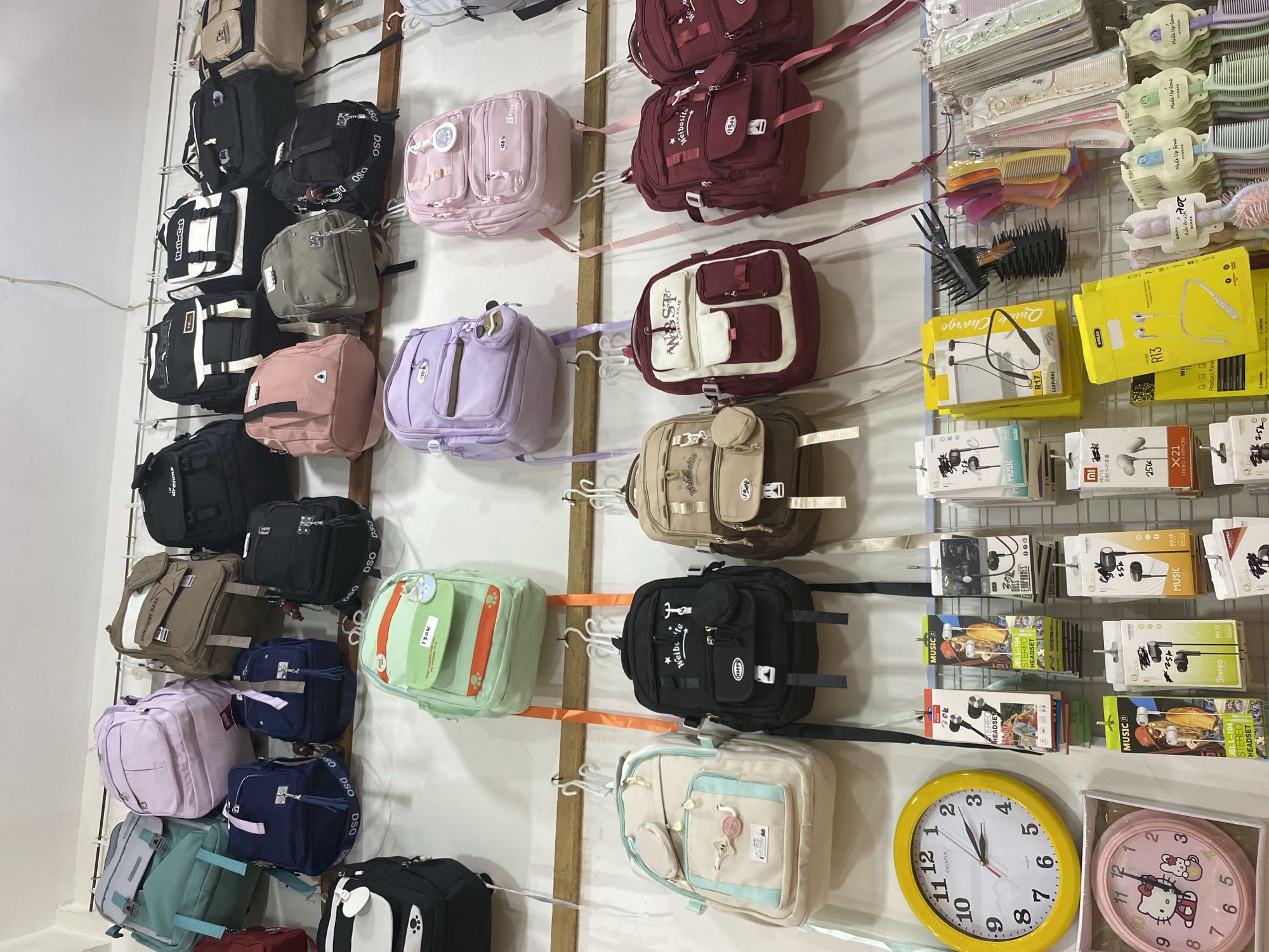 The image shows a wall display of backpacks and other accessories in a shop. The display is organized on multiple shelves and hanging racks. Backpacks of various colors, sizes, and styles are prominently featured, including pastel-colored options, dark-colored practical bags, and some with unique designs. Accessories like headphones, hair combs, and clocks are also visible, suggesting a retail environment that sells a variety of goods. One of the clocks is a yellow analog clock, while another is a pink circular clock with a Hello Kitty design. The overall impression is that of a well-stocked store, possibly catering to a younger demographic or those interested in trendy accessories. The store appears to be located in Lamteumen Barat, Indonesia, implying a local business serving the community.