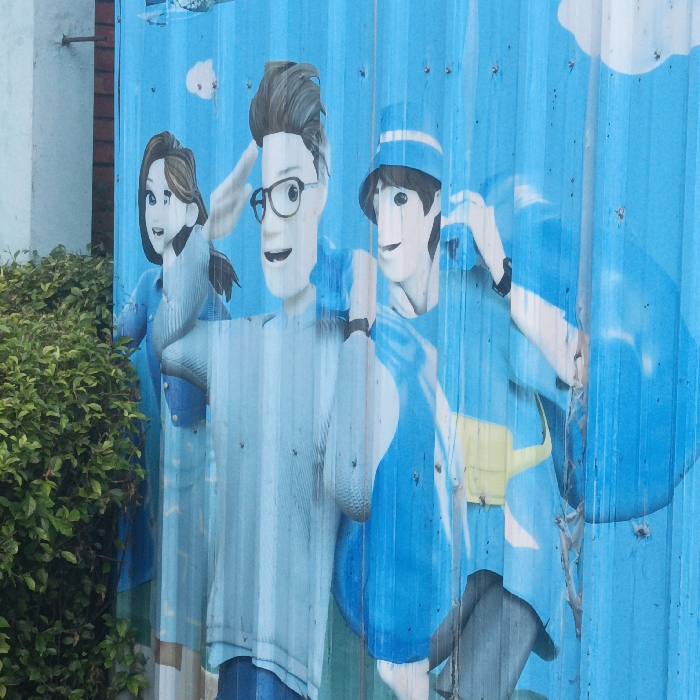 The image displays a brightly colored mural or large-format print affixed to a corrugated metal wall or fence, located within the Special Capital Region of Jakarta, Indonesia. The dominant color of the mural is a vibrant blue, serving as the background for three stylized, cartoon-like human figures.
From left to right:
1. A female figure with brown hair, depicted in a light blue long-sleeved top and a lighter blue lower garment. Her left arm is raised towards her head, and a dense green bush with small leaves partially obscures her lower body and the far left edge of the mural.
2. A male figure with dark, spiky hair and black-rimmed glasses, wearing a light blue long-sleeved shirt. His right hand is raised to his forehead, possibly in a salute or to shade his eyes, and he displays a wide, open-mouthed smile. A black wristband is visible on his left wrist.
3. A male figure wearing a light blue bucket hat and a bright blue long-sleeved top. His right arm is bent, and his hand is near his head, appearing to adjust an object. A black wristband is on his left wrist, and a yellow fanny pack or bag is secured around his waist. A large, lighter blue, indistinct shape, possibly a cloud or a large bag, is visible behind him.
The corrugated texture of the metal surface causes noticeable vertical undulations and slight distortion in the printed image. Fasteners, likely screws or rivets, are visible across the surface, securing the mural. Small, faint white cloud-like shapes are present in the upper portion of the blue background. To the far left, behind the bush, a segment of a light-colored building wall is discernible. The scene is illuminated by natural, presumably daylight, indicating an outdoor setting.
