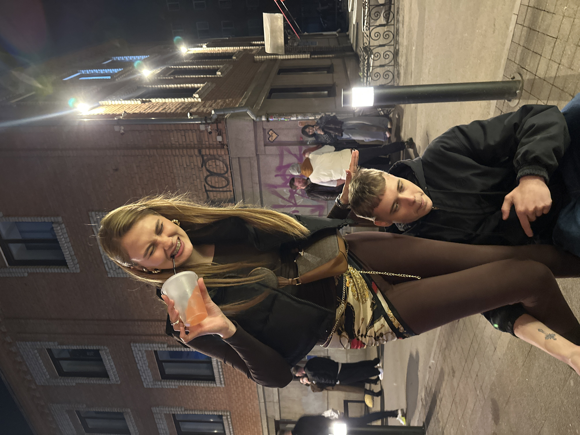 A medium shot depicts a young woman holding a drink with a straw in an outdoor setting. She has long blonde hair, smiles, and wears braces. She is dressed in dark clothing and accessorized with gold jewelry and a patterned purse. A young man in a black jacket is seen next to her; he is leaning back and looking towards the woman. They appear to be interacting, with the man possibly touching the woman's head.
The background features a brick building with windows, a street lamp, and a sidewalk. Graffiti is visible on the building's wall. Additional people are seen in the background, suggesting an urban setting. The photo was taken at night with artificial lighting. There is no definitive proof but based on graffiti and street view, the location is near Krasnoyarsk, Russia.