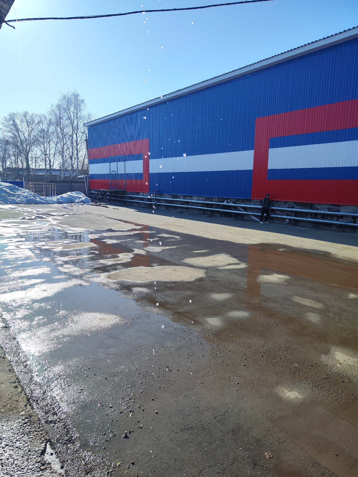 The image depicts a wet industrial area in Chaikovsky, Russia. A long, blue, red, and white striped building dominates the background, potentially a warehouse or factory. A pile of snow remains near some bare trees, suggesting it is early spring. In the foreground, the ground is wet with puddles reflecting the building and the sky. A person stands near the building, partially obscured by the shadow of the building itself. A metal guardrail runs along the length of the building. The overall impression is of a utilitarian, slightly desolate industrial landscape.