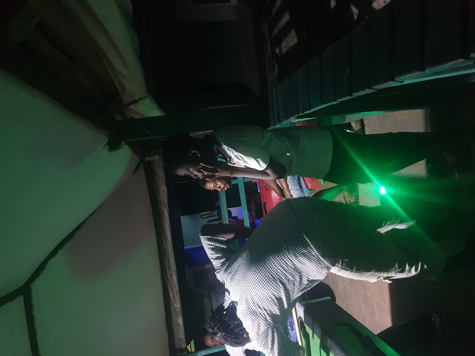 The image depicts a group of young men gathered at a dimly lit outdoor establishment, possibly a bar or lounge, in Lokoja, Nigeria. The scene is illuminated by green lighting, creating a moody ambiance. In the foreground, a person wearing a black and white patterned shirt and light-colored pants is bent over a pool table, appearing to be engaged in a game. The pool table is green and has cues and balls visible. To the right of this individual, another young man, wearing a light-colored polo shirt, is standing and looking away from the camera with his hand near his mouth, deep in thought. Further in the background, other individuals are partially visible, some seated and others standing. The environment suggests a casual recreational setting, with wooden structures and benches visible in the background, illuminated by the green lights and the dim glow of the night. The overall impression is one of a social gathering enjoying a game of pool in an outdoor Lokoja nightspot.