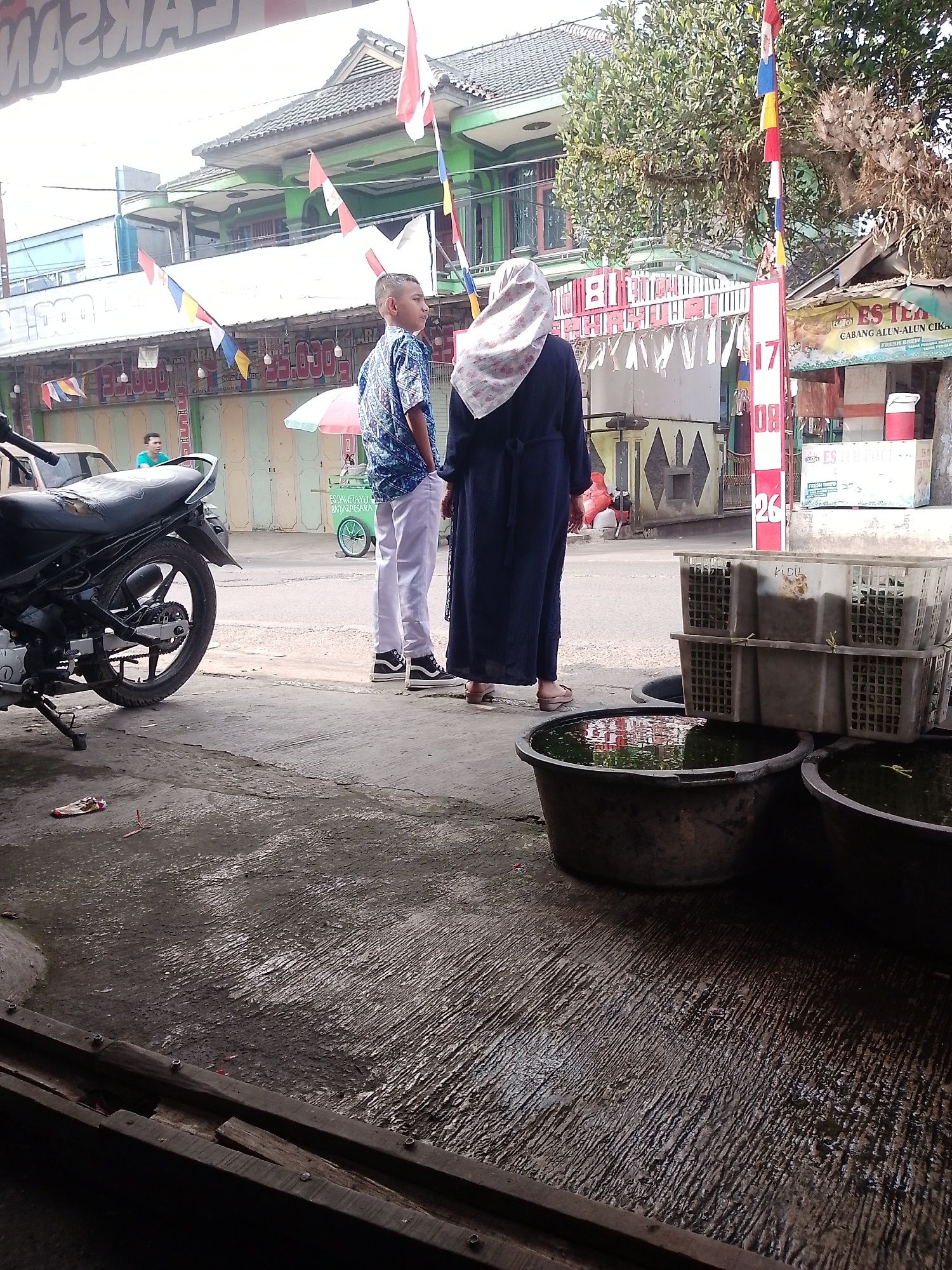 In Cikalongkulon, Indonesia, a street scene unfolds near a building with a green facade and a tiled roof. In the foreground, a dark motorcycle is parked on a concrete surface. To the right of the motorcycle, two individuals stand facing away from the camera. The person on the left is a young male wearing a blue and white patterned shirt and white pants. The person on the right is an adult wearing a dark blue abaya and a light-colored hijab. They are standing on a paved area. Several large, dark circular tubs, some filled with water, are positioned in the foreground to the right. Behind the individuals, a green cart with a pink umbrella is visible. Further back, a building with a white facade and various banners and flags is present. A small red and white sign with text is also visible on the right side of the image. The overall environment appears to be an outdoor marketplace or street setting with commercial establishments.