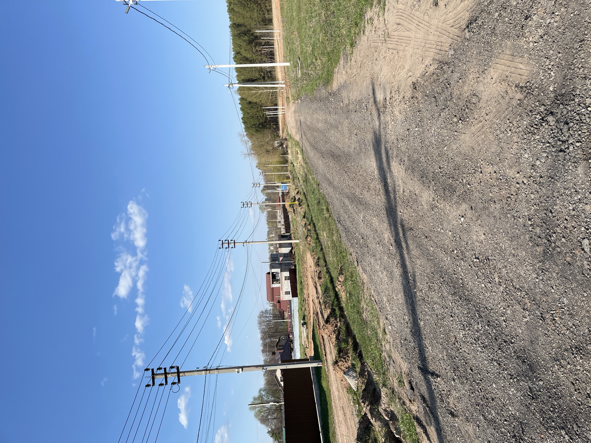 The image shows a dirt road in a rural area. Power lines run alongside the road, supported by poles. Several houses and buildings are visible in the background. A forest and a field are also visible. The location is Chaikovsky, Russia.
