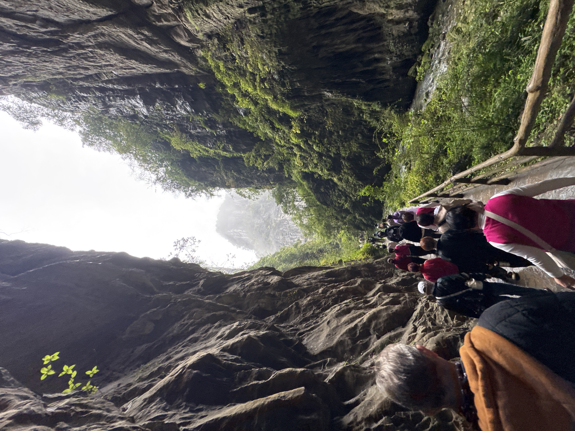An outdoor shot of a group of people on a hiking trail along a canyon wall. The photo is taken looking up the canyon wall, and the angle is askew, with the hikers on the right. The canyon walls are on either side of the group, with a bright white sky visible in the distance. The rock face on the left of the image is a dark grey. Bright green foliage is scattered across the rock face on the right. A wooden railing sits to the right of the hikers. This location is in the city of Wulong District, China.