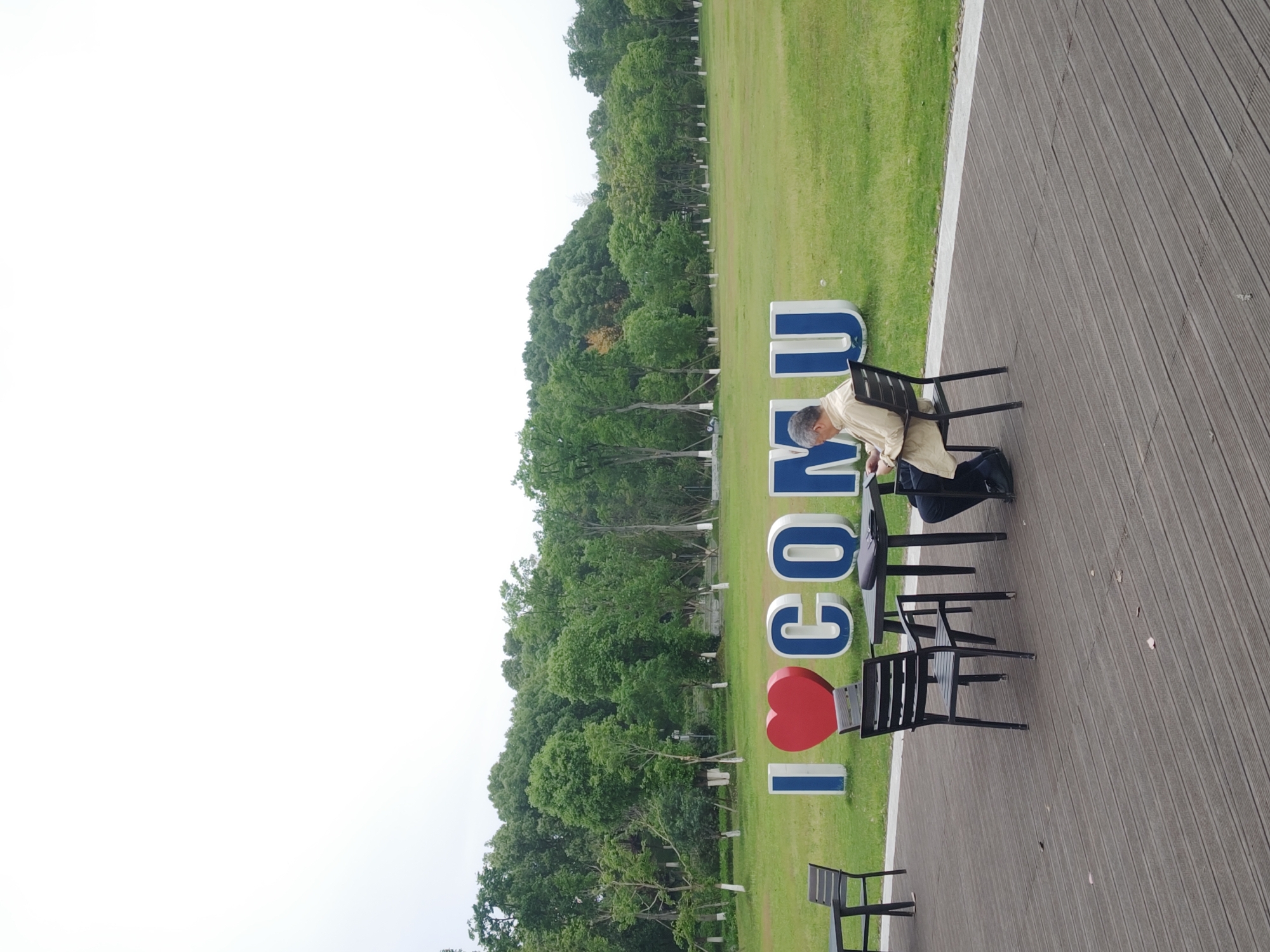 The image shows an outdoor scene in Chongqing High-tech Zone, China. A man sits on a black chair, leaning slightly forward. Next to him are two empty black chairs, and another chair is positioned nearby. The background features a grassy area with trees and a partially visible sky. To the left of the man are large, vertical letters and a heart symbol spelling out "I <3 CQMU". The chairs and the man are on a wooden deck.