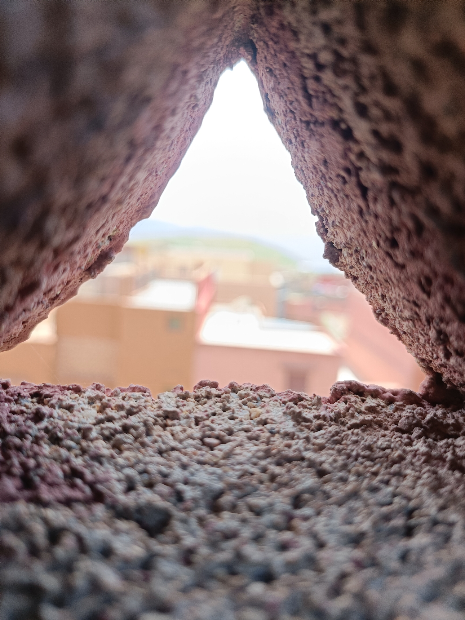 The image shows a close-up view through a triangular opening in a rough, reddish-brown stone or brick wall. The wall texture appears porous and coarse. The background visible through the opening is a blurred view of a town, presumably Ouaouizert, Morocco, with buildings in shades of tan and orange. The background also includes a faint green hill or field under a bright, hazy sky. There are no people visible.