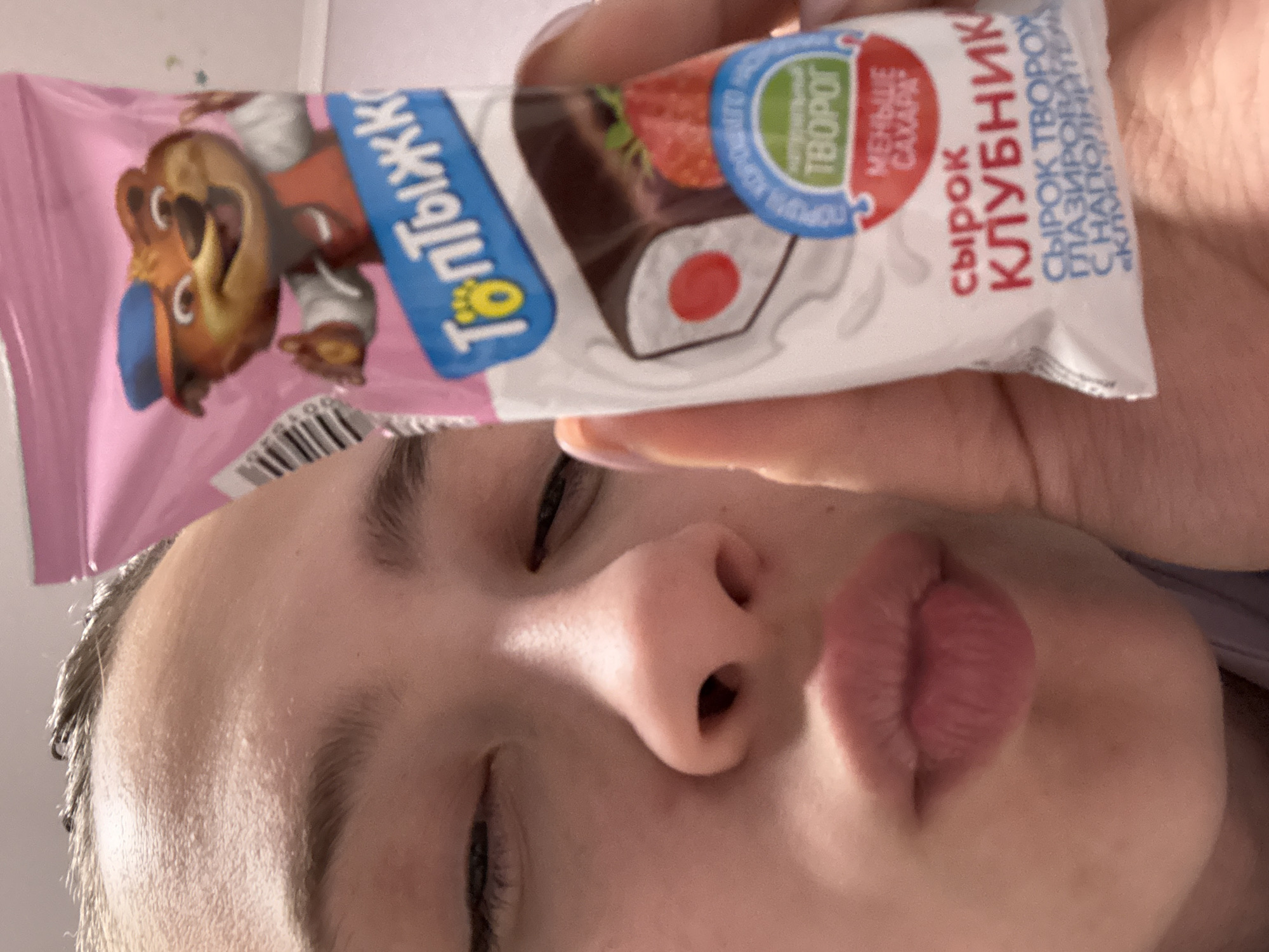 A person is holding a Toptyzhka brand strawberry-flavored cottage cheese bar in front of their face. The bar has two wrappers: a pink wrapper with a cartoon bear and a white wrapper with an image of the snack and Russian text. The text on the wrapper includes "СЫРОК КЛУБНИК," "Творог" (cottage cheese) and "Меньше сахара" (less sugar). The location is the city of Прикамский, Russia based on the language of the packaging.