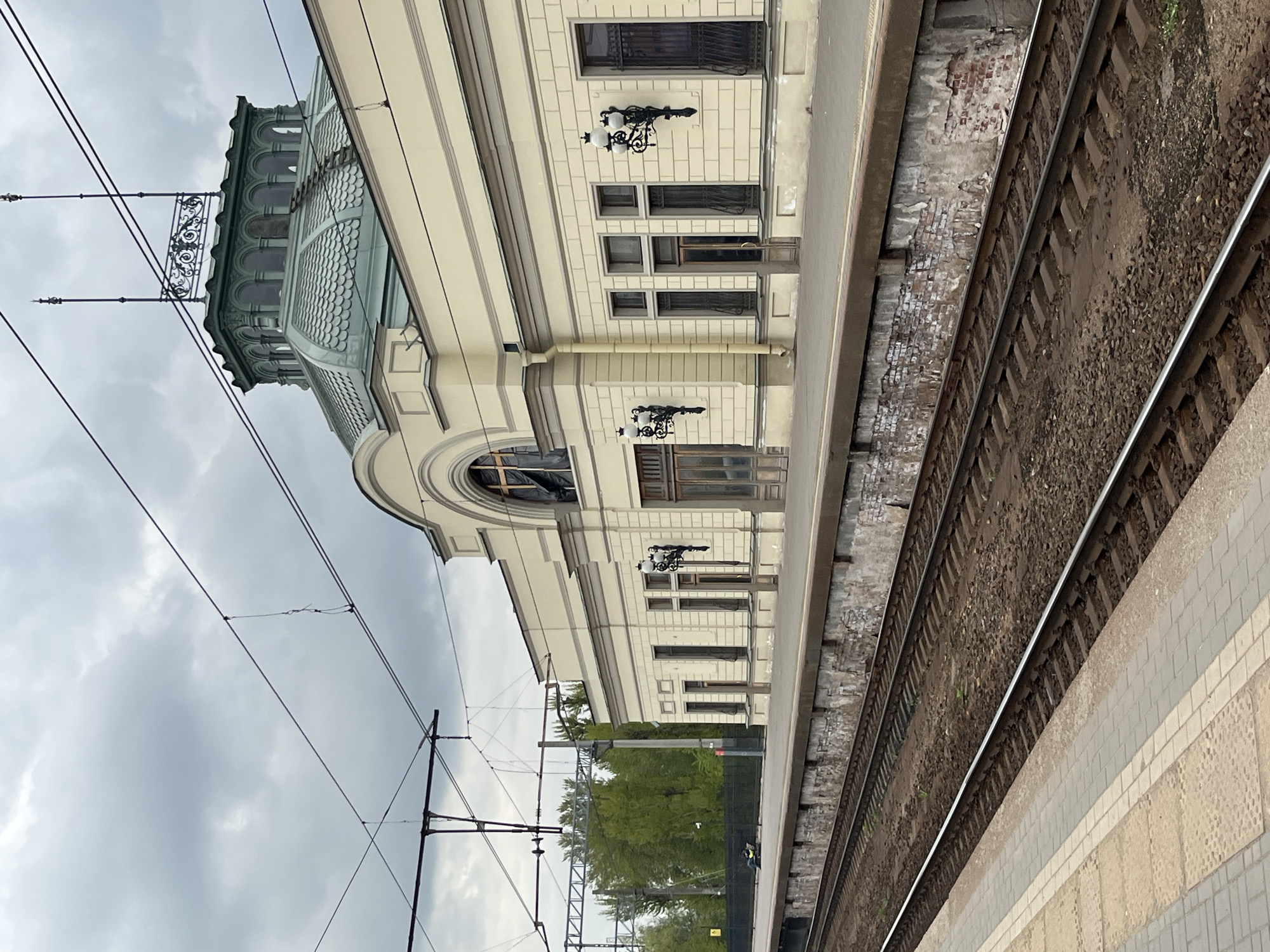 The image shows a train station with a building next to the tracks. The building is light yellow with black wrought-iron lamps on the exterior. It has a rounded arch with an arched window. The building has a turret or cupola with a filigree railing. In the foreground are train tracks with brown gravel between the rails. Next to the tracks is a tiled platform. Overhead are electrical lines for the train. In the background, there is green foliage. The sky is gray and overcast. The location is Unknown, Unknown.
