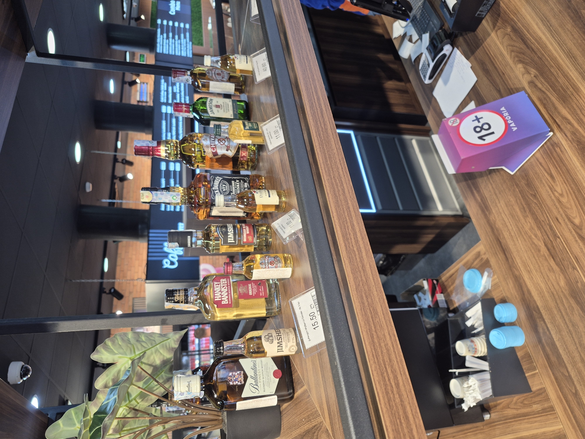 The image shows a display of alcoholic beverages on a wooden shelf, likely inside a store in Berbuki, Georgia. The focus is on various bottles of whiskey, including brands like "Ballantine's", "Jim Sher", "Hankey Bannister", "Jack Daniel's" and "Chivas Regal". Price tags are visible next to some of the bottles. A small indoor plant sits at the front of the display. In the background, there is a cash register and a sign with "18+" and "Vaporia" branding. A stack of disposable cups is also visible. The overall environment suggests a retail setting, possibly a supermarket or liquor store.