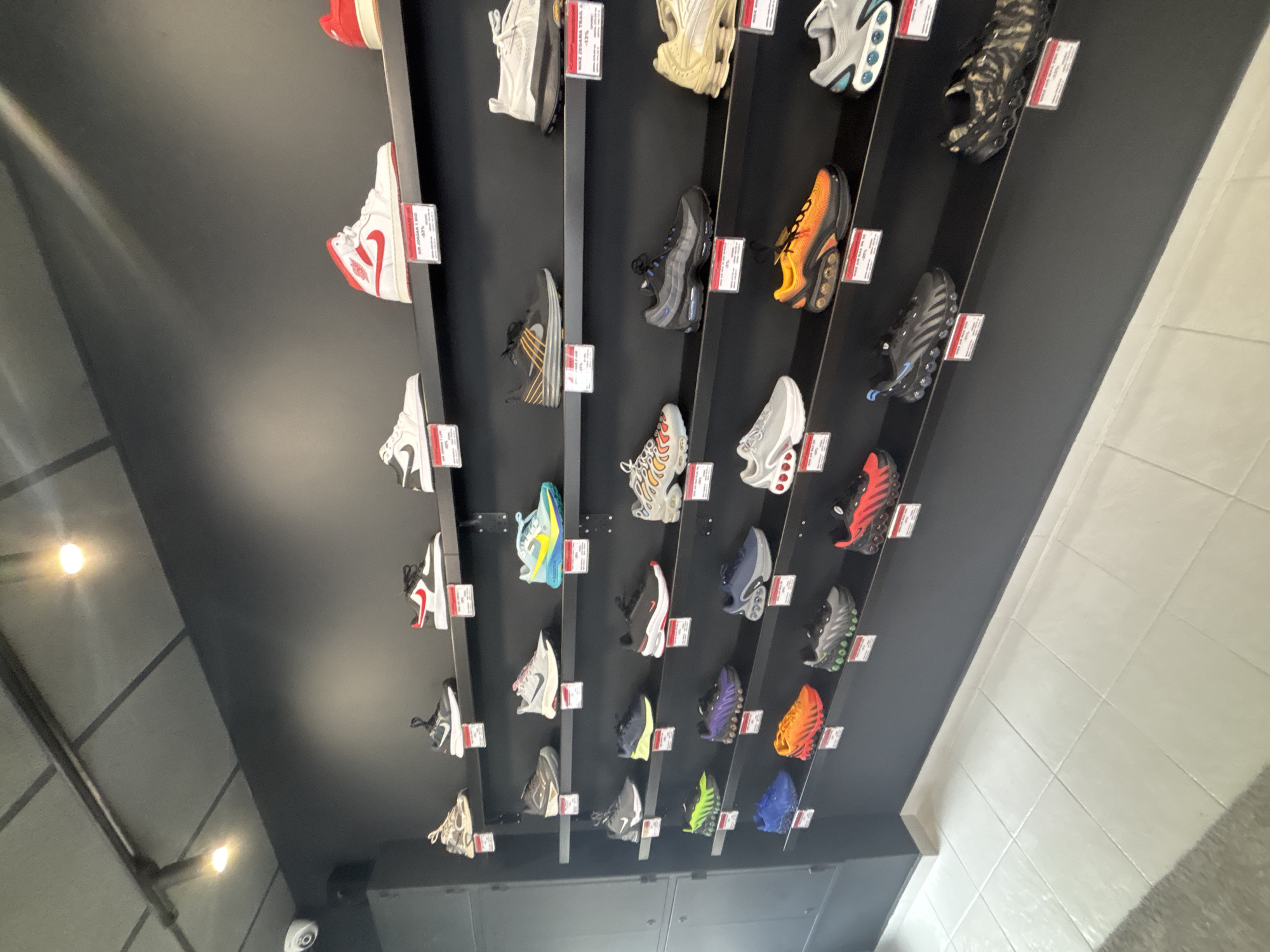 The image shows a display of sneakers on a wall in a store. The sneakers are arranged in rows on vertical metal bars. Each sneaker has a small tag attached to it. The wall behind the sneakers is black, and the floor is visible at the bottom of the frame. The environment looks like a retail shoe store and it is located in Gabrovo, Bulgaria.
