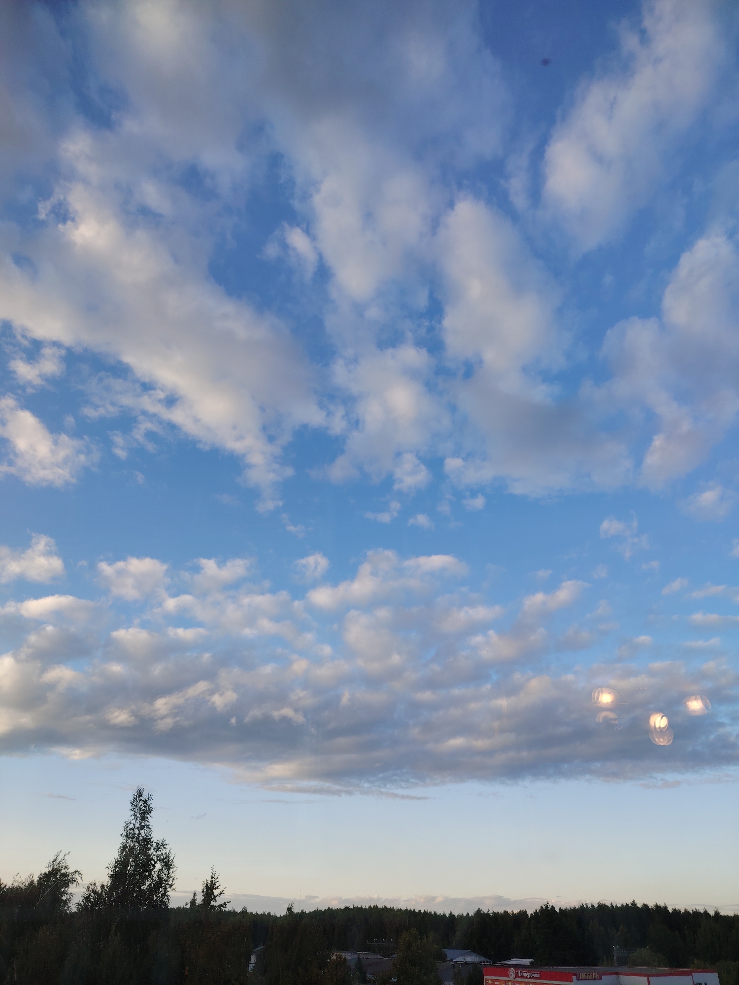 The image shows a cloudy sky over a treeline with some buildings visible in the distance. The sky is filled with white and gray cumulus clouds against a blue background. Below the sky, a dark green band of trees stretches across the horizon. Some scattered buildings are peeking through the trees. One building has a red sign with white lettering that appears to say "Пятёрочка" and "МЕБЕЛЬ", which are Russian words for a supermarket chain and furniture store, respectively. The scene is likely a view from a slightly elevated position looking towards the edge of the city of Konakovo, Russia. There are no people or specific actions depicted in the image. The overall impression is a late afternoon or early evening scene with diffused sunlight.