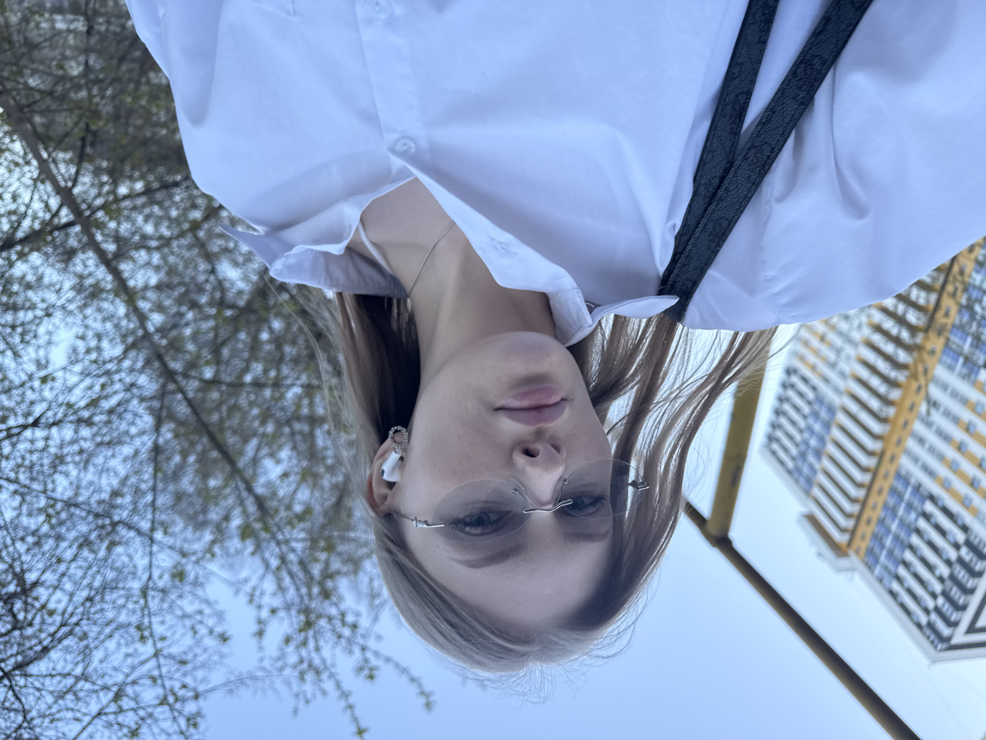 A woman with light brown hair and light eyes, wearing rimless glasses and a white collared shirt, appears to be taking a selfie. She has an earbud in her left ear and a dark-colored strap hangs over her shoulder. The background shows a blurred view of trees with budding leaves and a tall, modern apartment building with yellow accents against the blue sky. Based on the architectural style of the building, the photo was taken in Perm, Russia.