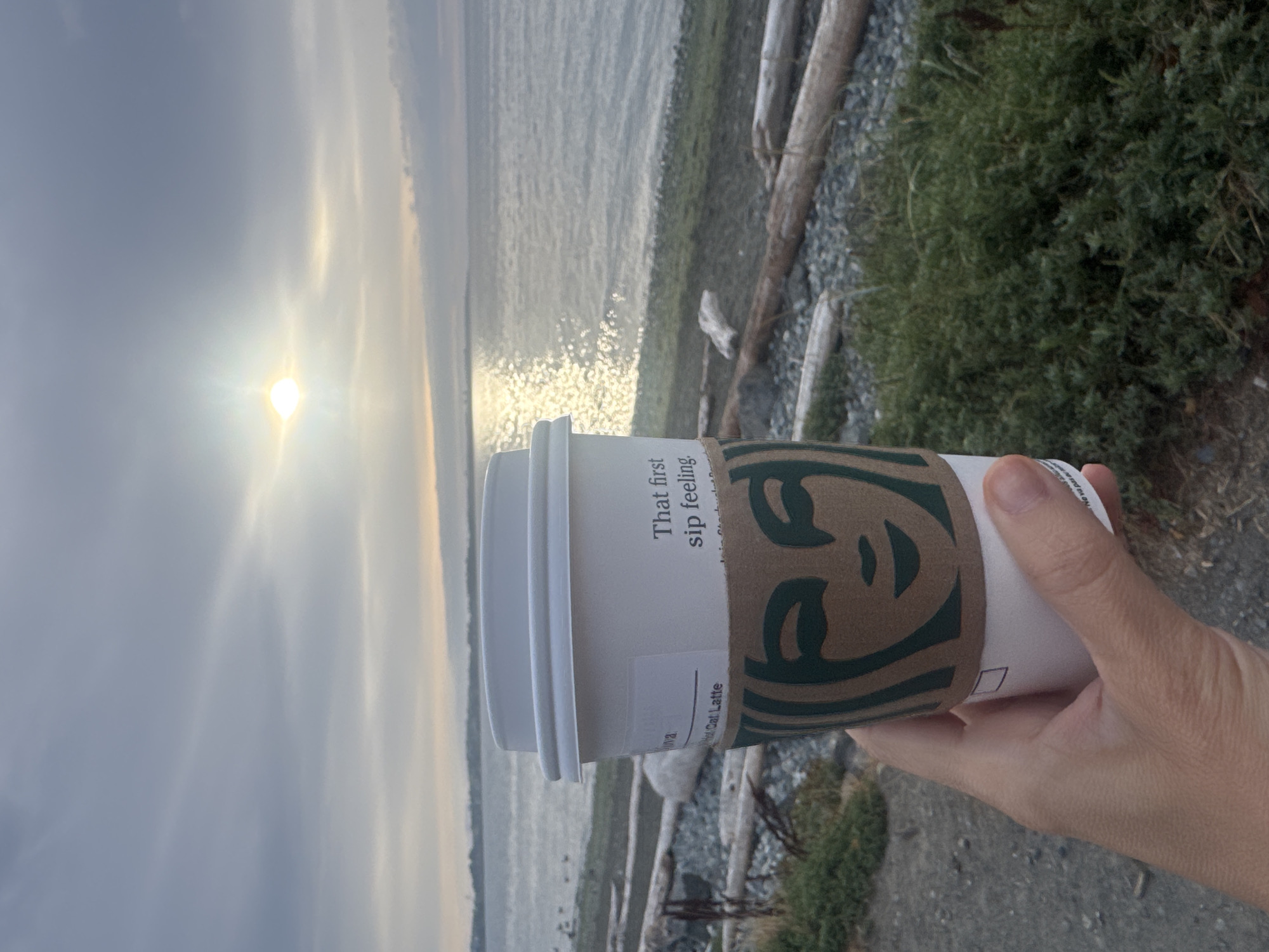 A person holds a white Starbucks coffee cup with a brown cardboard sleeve featuring the Siren logo. The cup is positioned in the foreground, slightly to the left of center, with a hand visible holding it. Behind the cup, a body of water, likely the ocean, stretches to the horizon. The sun is setting or rising, casting a bright glare on the water and clouds in the sky. The shoreline is visible, composed of pebbles and driftwood. Green vegetation is present on the land. The scene appears to be a coastal area. This image was captured in Colwood, Canada.