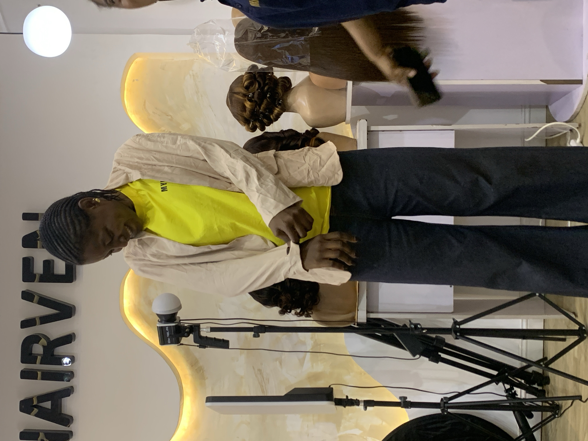 The image captures an indoor scene, likely a hair salon, beauty studio, or retail space in Animashawun, Nigeria. The central subject is a person, appearing to be a young individual, standing and looking downwards. They have dark skin and braided hair, wearing a bright yellow t-shirt with "MY VE" partially visible text, and a light beige or cream-colored long-sleeved shirt or jacket over it, paired with dark trousers.
In the background, there are modern, curved display units or shelves, illuminated from behind with a warm yellow light. Several mannequin heads are visible on these displays, showcasing various wigs or hair extensions. One wig is styled with prominent curls, while another is covered in clear plastic wrapping.
To the left of the central figure, prominent large, dark letters spell out "HAIRVE" against a white wall, suggesting the name of the establishment or brand. In the foreground, photography or videography equipment is set up, including a black tripod stand holding a long, rectangular LED panel light and another smaller, spherical light source.
Towards the top right of the image, a hand belonging to another partially visible person is seen holding a mobile phone, possibly filming or taking a picture. A bright white circular light fixture hangs from the ceiling. The overall setting appears to be designed for showcasing hair products or services, possibly during a promotional event or photoshoot.