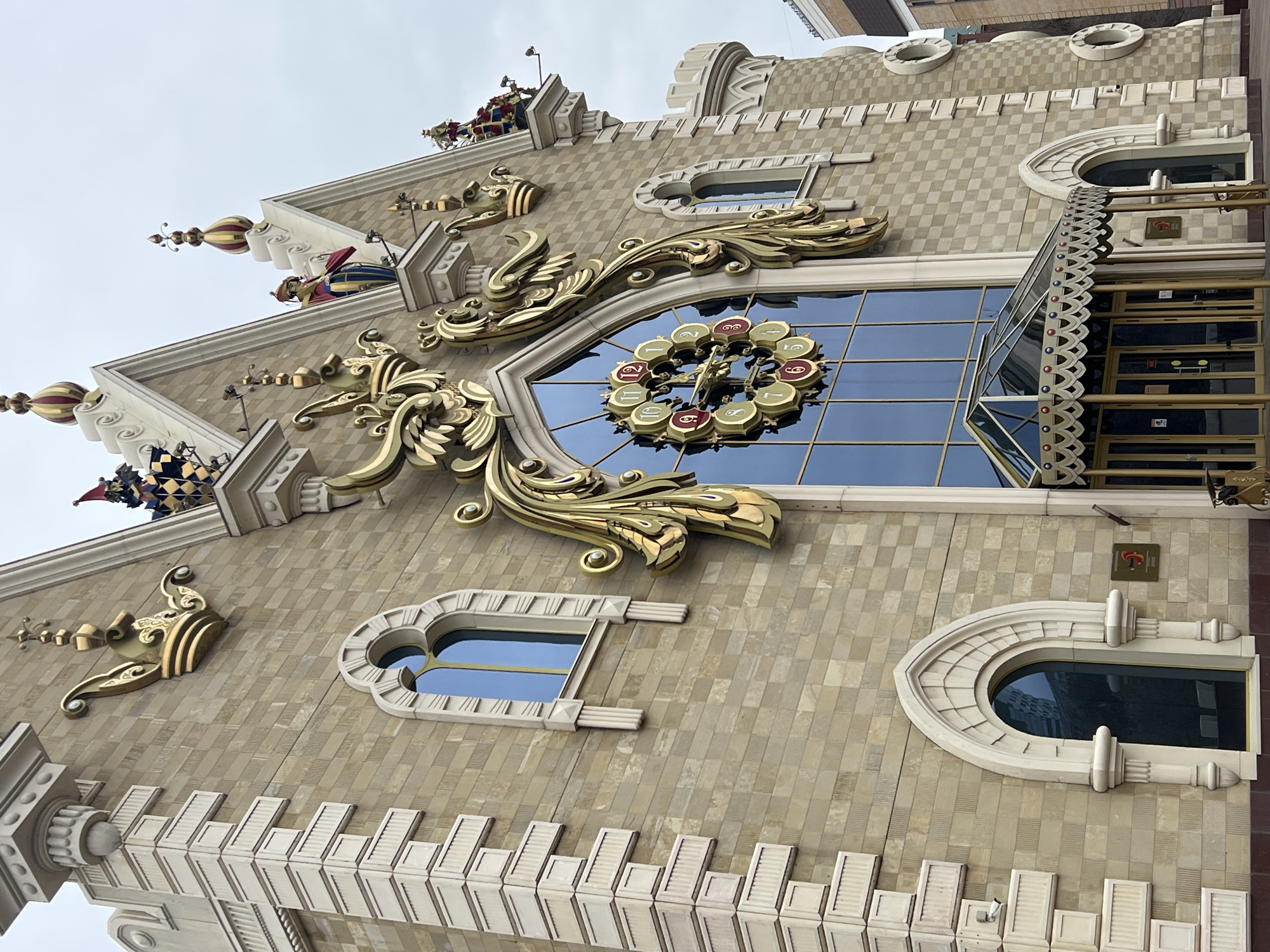 The image depicts a building facade in Kazan, Russia, recognizable by its ornate, fairy tale-like architecture. The building is constructed of light-colored, brick-like blocks arranged in a checkered pattern. The facade features decorative golden embellishments, including scrollwork and a large clock with oversized numerals. The building has arched windows of varying sizes and shapes. At the top of the building are ornate figures, and spires with decorative elements. To the right of the clock is what appears to be the entrance, protected by a canopy and railings. The sky is overcast, suggesting a cloudy day. The architectural style is reminiscent of fairytale castles, making it a distinctive landmark in Kazan.