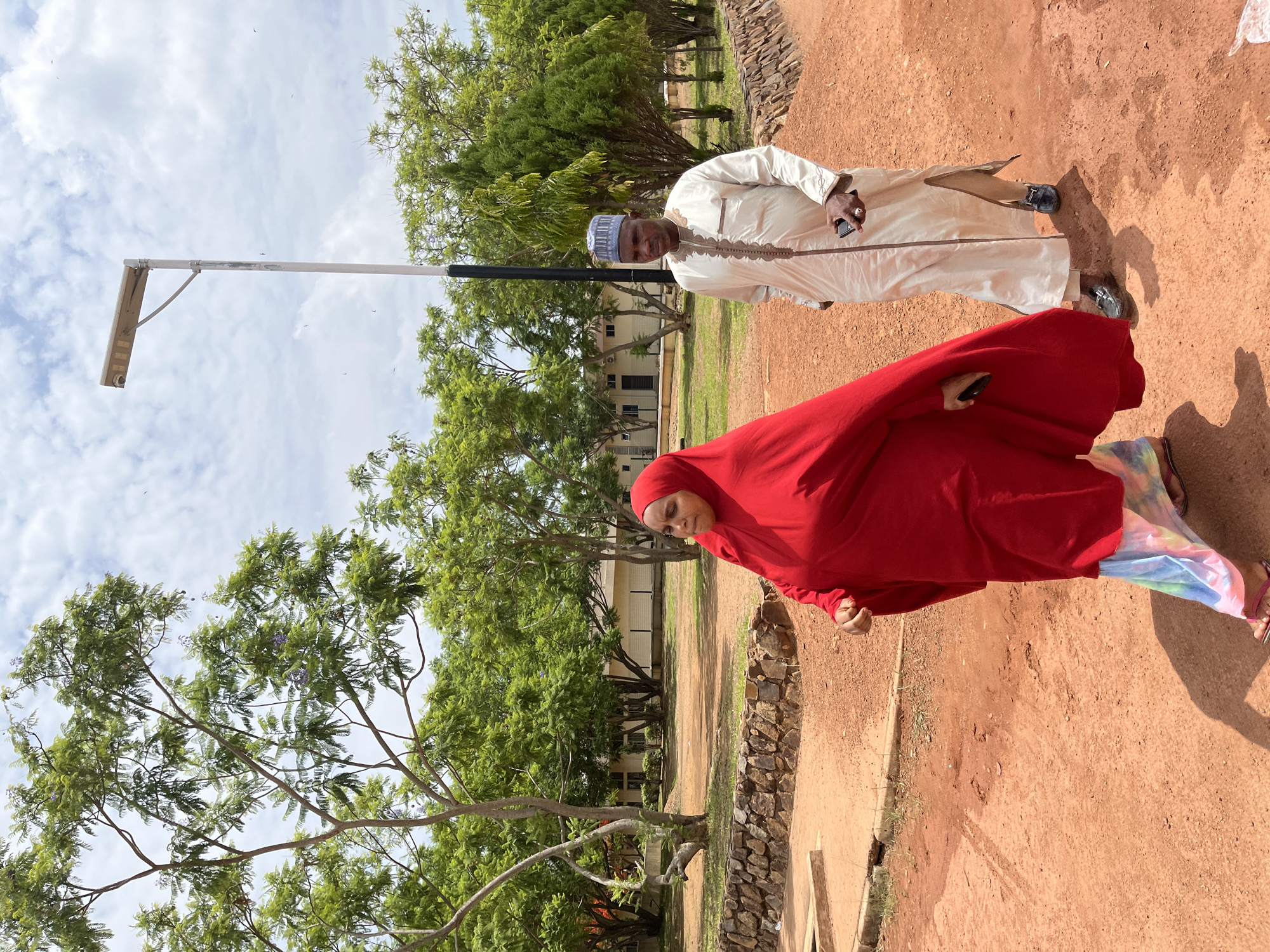 Two people stand on a dirt and grass area in Gwong, Nigeria. One person, wearing a red hijab, is positioned in the foreground. The other, an older man wearing a white robe and a blue and white cap, stands behind her. Both are holding phones. A metal pole with a light fixture is located to their left. Trees and a building are visible in the background. A low rock wall separates the dirt area from a grassy section. The sky is partly cloudy.