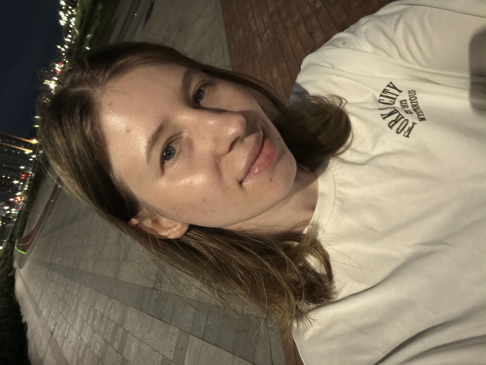 In this image, a young woman with shoulder-length brown hair and blue eyes is taking a selfie. She is wearing a white t-shirt with a logo that reads "FORKS CITY EST 1873 MYSTERIOUS". The background shows a paved walkway, likely in a park or city area, with lampposts and distant city lights visible, suggesting it is nighttime. The location is identified as Dongducheon-si, South Korea. The woman is looking directly at the camera with a slight smile. The lighting suggests artificial light sources.