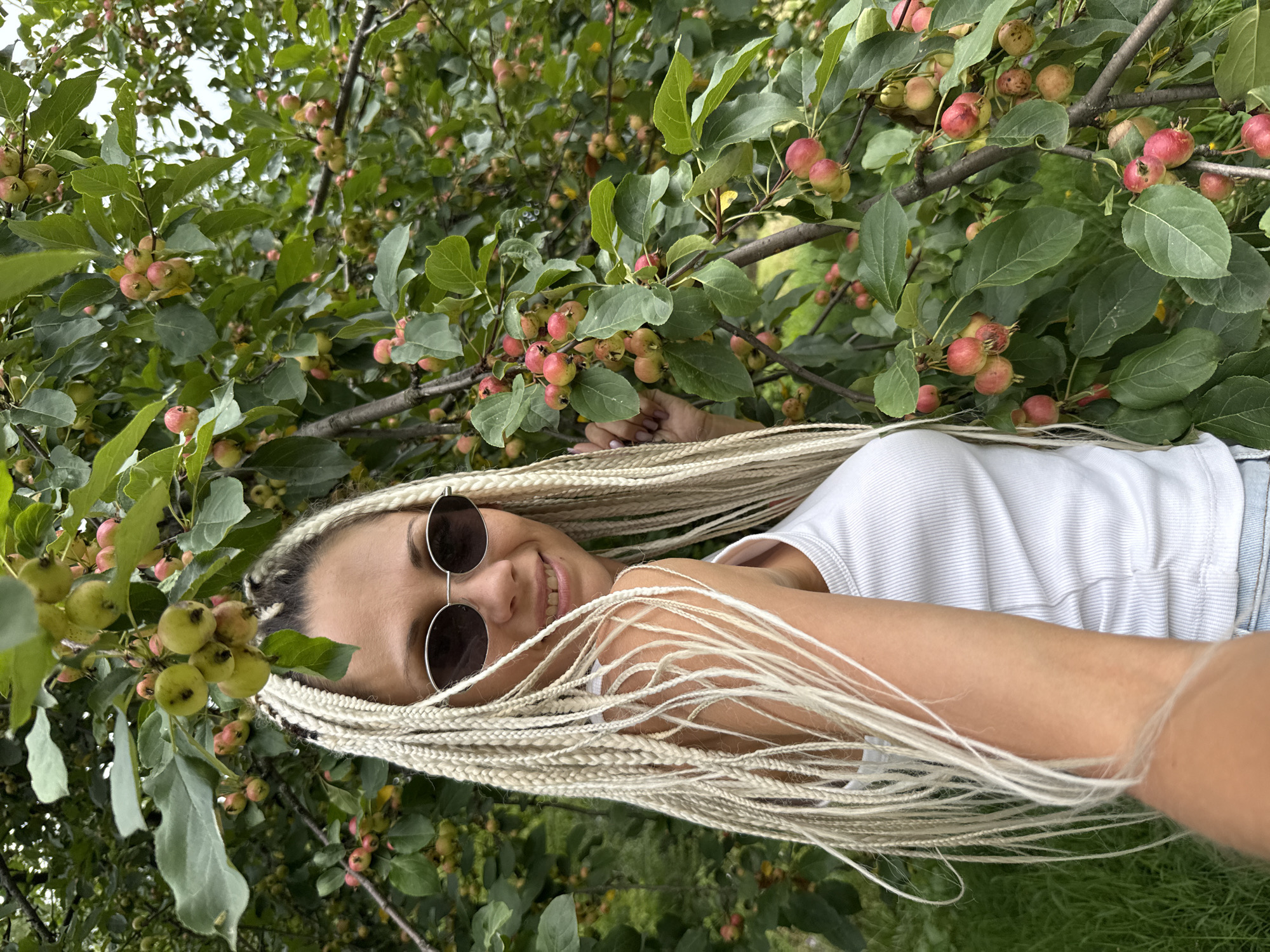 A blonde woman with long braids and sunglasses stands in front of a tree with small red and green fruits. She is wearing a white tank top and denim shorts. The background is filled with green leaves and branches. The scene is set outdoors, likely in a natural setting. The location is specified as Rusanovo, Russia.