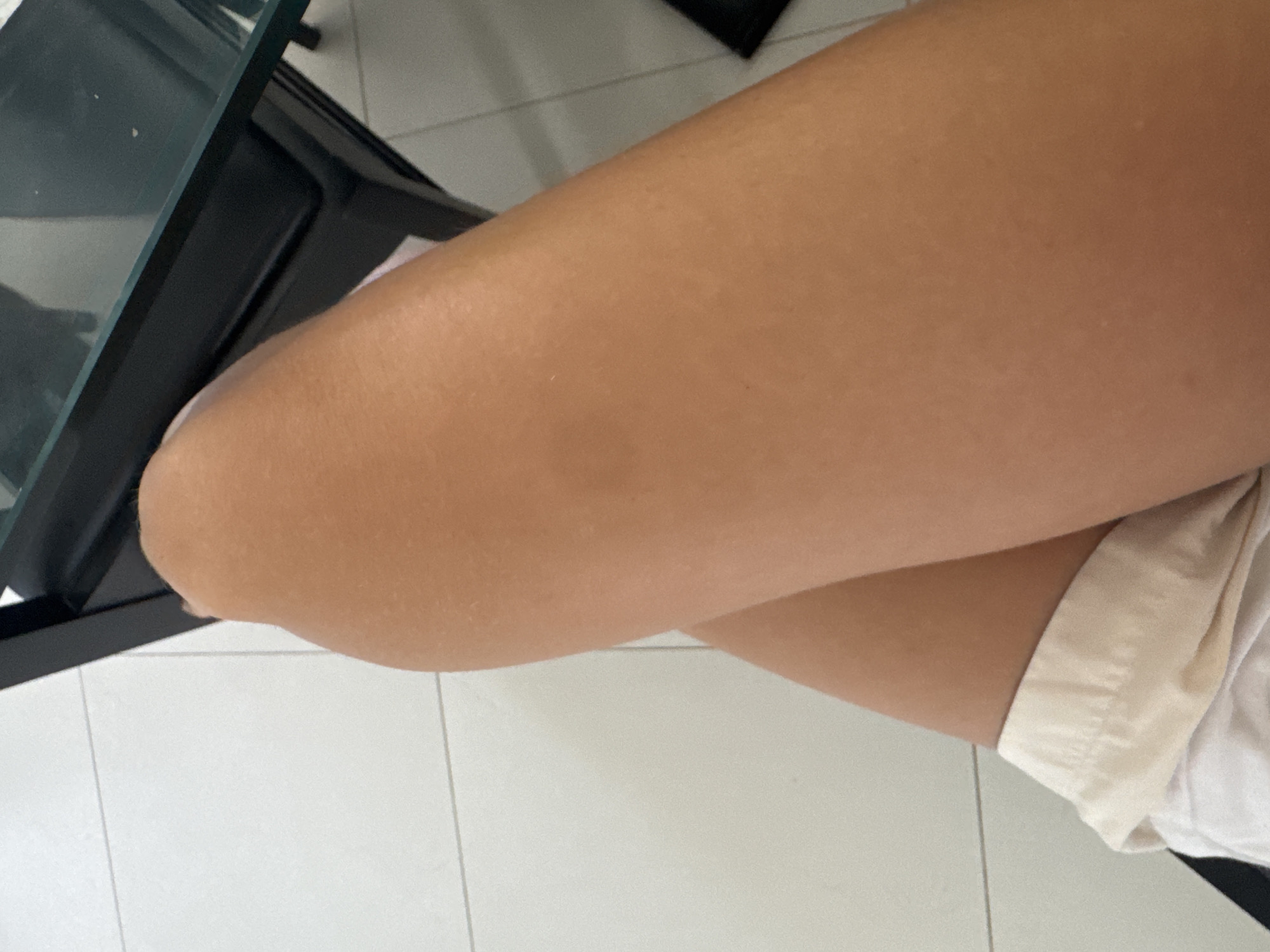 The image shows a close-up view of a person's legs, with their knees crossed, in what appears to be an indoor setting. The legs are tanned and bare from the knee down, with white fabric visible at the bottom, likely shorts. In the background, a portion of a dark-colored table with a glass top is visible, and a tiled floor is also present. The lighting suggests it is daytime. The scene is likely in a public space or a private residence, and given the context of Ko Samui District, Thailand, it could be a hotel room, a cafe, or a beachside villa.