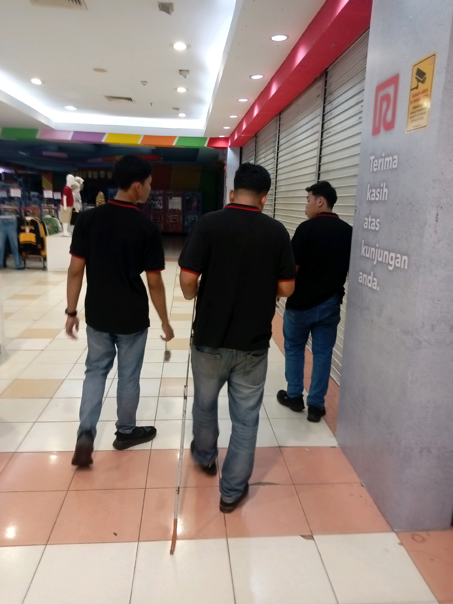 Three individuals, appearing to be male, walk down a brightly lit indoor mall corridor in Batam, Indonesia. The two individuals in the foreground are wearing black polo shirts with red trim and blue jeans. The person furthest to the left has a black watch on their left wrist and is holding a white cane. The person in the middle appears to be walking slightly ahead and to the right of the first individual, also holding the white cane, which extends to the floor in front of them. The third individual, further to the right and walking behind them, is also wearing a black polo shirt and blue jeans. The wall on the right side of the corridor has a large red logo and text in white that reads "Terima kasih atas kunjungan anda" (Thank you for your visit). The floor is tiled with a pattern of pale pink and white squares. In the background, to the left, clothing racks and mannequins are visible, suggesting this is a retail area within the mall. The ceiling is white and illuminated by recessed lighting and cove lighting along the edges.