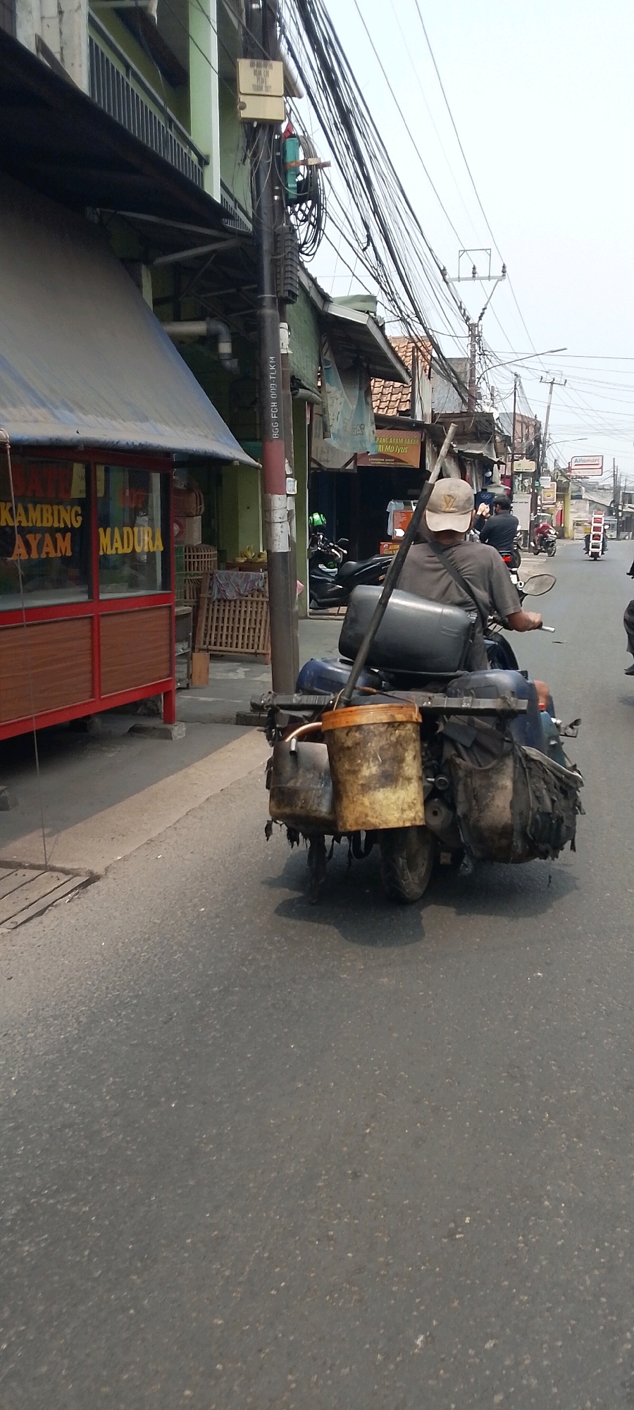 The image shows a man riding a motorcycle on a street in Bekasi, Indonesia. The man is wearing a light-colored cap and a gray t-shirt. He is carrying several items on the back of his motorcycle, including buckets and bags. In the background, there are buildings, utility poles with many wires, and other motorcycles. One building on the left appears to be a food stall with signage in Indonesian. The street is paved and appears to be a typical urban environment. The sky is overcast.