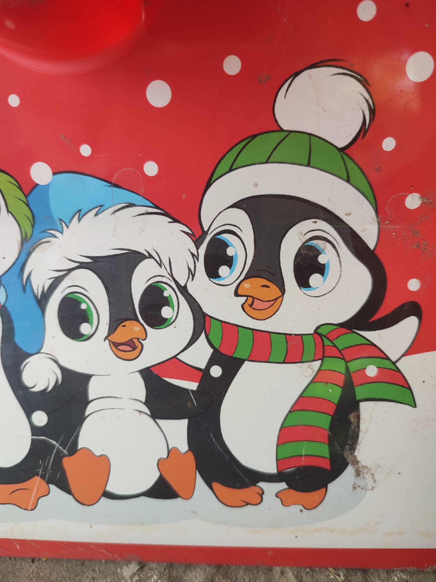 This is a close-up, eye-level, outdoor shot of a close-up of a drawing of two cartoon penguins. The drawing is on a red surface, possibly metal, with scattered white circles representing snow or confetti. The penguin on the left is wearing a blue winter hat with a white fluffy trim and has green eyes. It is looking towards the right with a slight smile. The penguin on the right is wearing a green and white striped hat with a white pom-pom and a red and green striped scarf. It has blue eyes and is also looking towards the right, with its beak open slightly and a smile. Both penguins are depicted in a cute, stylized manner with large heads and eyes. The drawing appears to be part of a larger design, possibly a decoration or a children's product. The overall impression is festive and cheerful, evoking a winter or holiday theme. There is no visible text in the image. The lighting suggests it is daytime, and the image is slightly dusty, indicating it might be an older item or has been stored outdoors. The location context is Shyshaky, Ukraine.