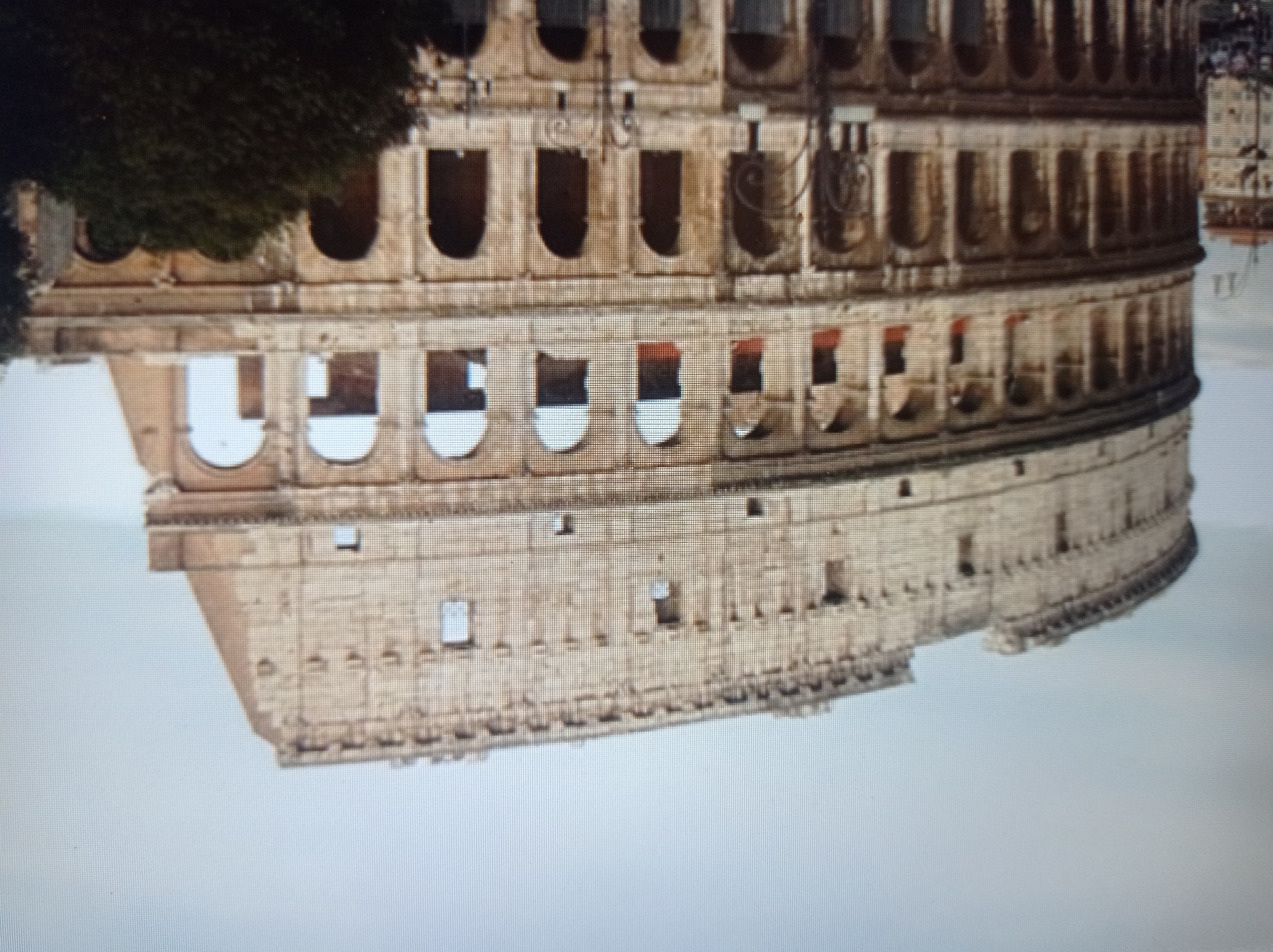 This is a photograph of the Colosseum in Rome, Italy. The massive, ancient amphitheater is made of stone and has multiple levels of arches. The sky above is overcast and gray. The image is slightly blurry and appears to be taken at an angle, capturing the structure from its side. In the foreground on the right, a dark green tree obscures a portion of the Colosseum. To the left, a street lamp is partially visible. The bottom of the image shows what appears to be a street with some indistinct figures and buildings. The location is clearly Rome, Italy, and not Mukachevo, Ukraine.