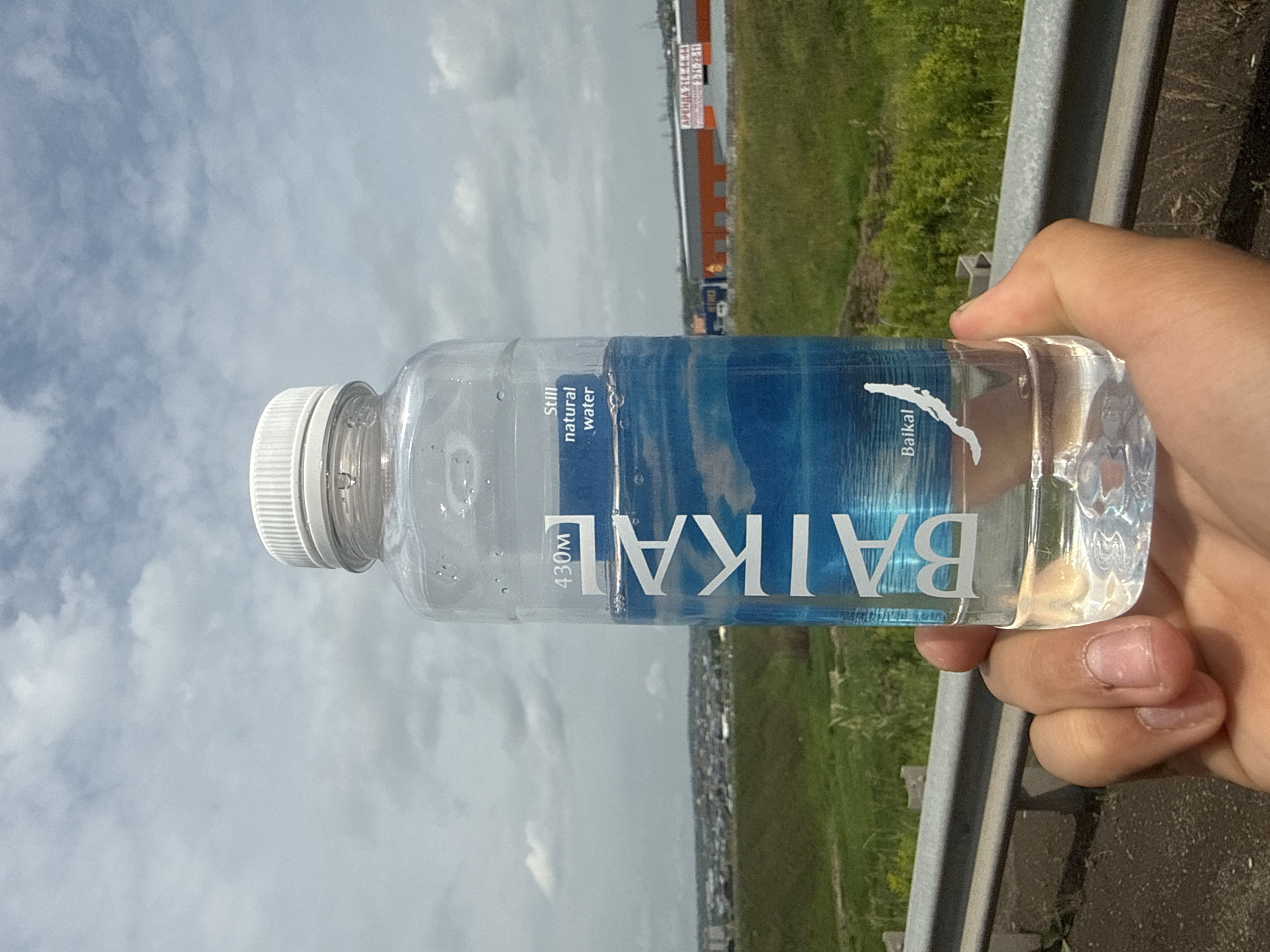 A person's hand is holding a clear plastic bottle of Baikal water. The bottle has a white cap and a blue label with white text. The label reads "BAIKAL", "430M", and "Still natural water". A silhouette of Lake Baikal is also visible on the label. The bottle is in focus, and the background is blurred. The background shows a cloudy sky, green grass, some buildings, and a highway guardrail. The scene appears to be outdoors, possibly in a suburban or rural area near the city of Solontsy, Russia, given the presence of industrial buildings and the clear sky. The image is taken from a low angle, looking up at the bottle and the sky.