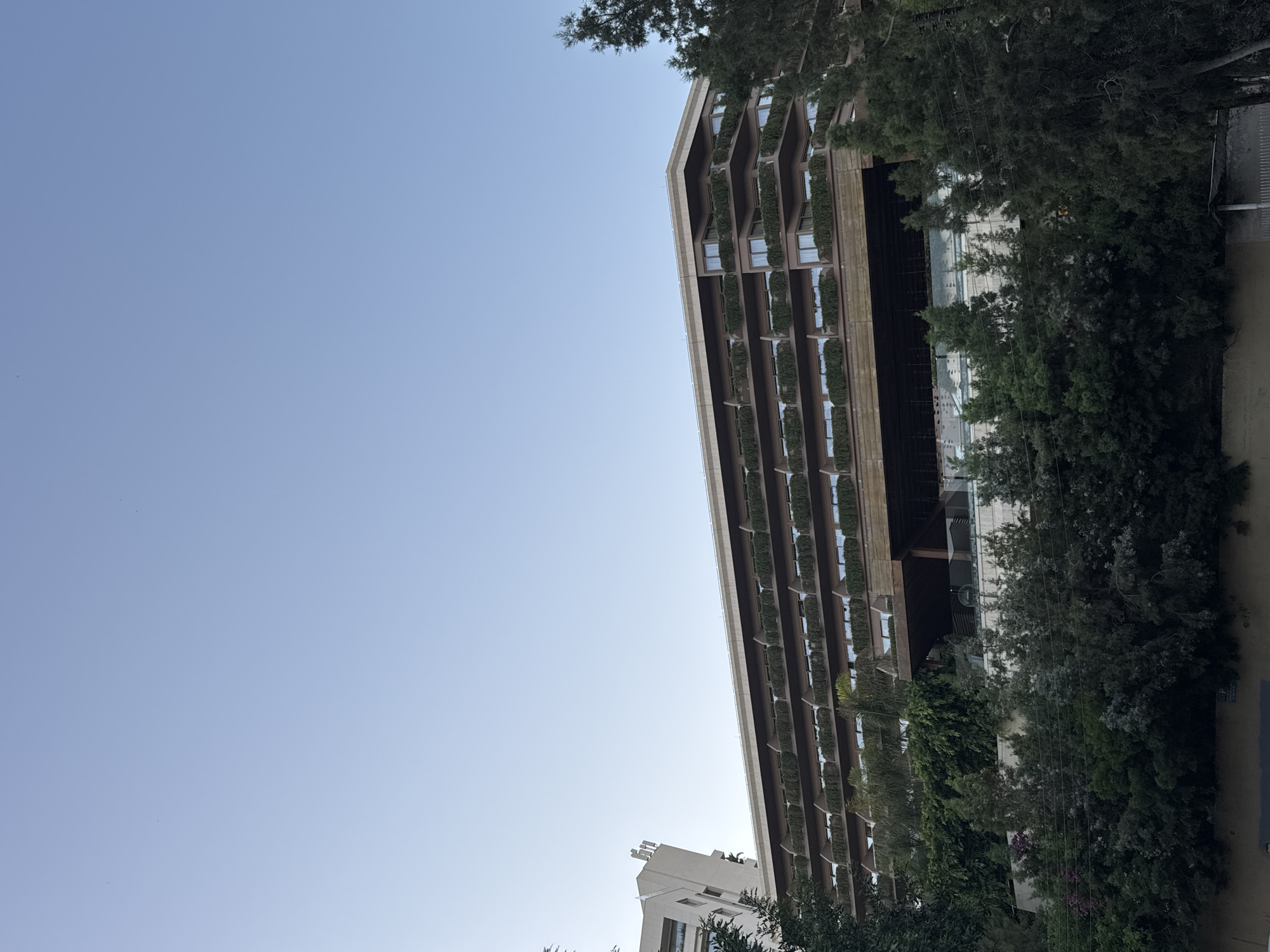 This is a low-angle exterior shot of a large, multi-story building in Agios Tychona Community, Cyprus. The building has a tiered, modern design with numerous balconies, each lined with lush green plants. The balconies are stacked horizontally, creating a layered effect on the facade. The main structure of the building appears to be clad in a warm-toned material, possibly wood or a textured composite, with windows visible behind the greenery.
To the left of the main building, a portion of another, lighter-colored building is visible. It has a sharper, more angular design and a rooftop sign with abstract lettering.
The foreground is dominated by dense foliage and trees, with some of the branches reaching up towards the lower levels of the buildings. The trees vary in size and density, creating a natural screen in front of the lower sections of the structures. Some power lines are visible running horizontally across the image, interspersed with the trees.
The sky above is a clear, pale blue, with no clouds present. The overall lighting suggests it is daytime, possibly late afternoon or early morning, given the soft light and absence of harsh shadows. There are no people or discernible activities visible in the image. The scene depicts a blend of modern architecture and abundant greenery.