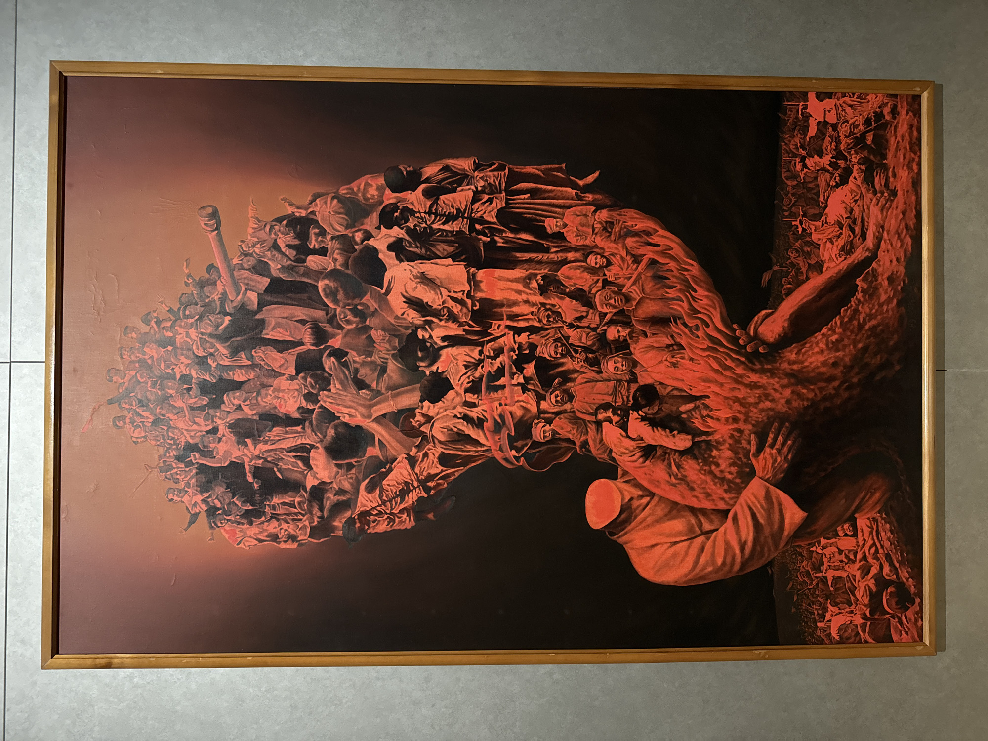 The image shows a framed painting with a dark red color scheme. The painting depicts a surreal scene with a large mass of people clustered together towards the top left, fading into a dark background. A figure in a white robe and no head is on the bottom right, holding onto what appears to be a rocky or mountainous terrain. The bottom right edge of the painting also features a dense collection of smaller figures, possibly representing a crowd. The painting is hanging on a wall, possibly in an art gallery. The painting is located in the city of Seoul, South Korea.