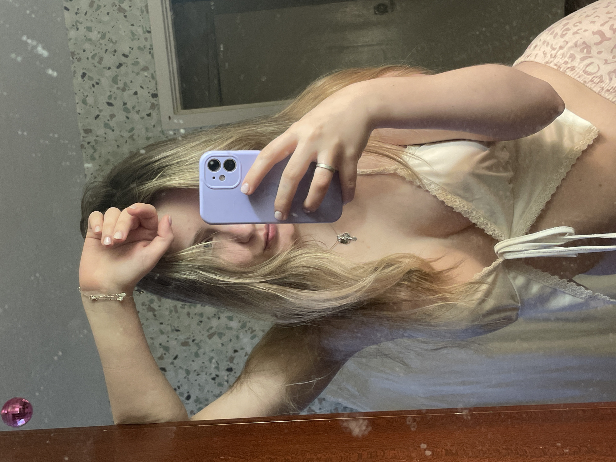 A young woman with blonde hair takes a mirror selfie in a bathroom. She is wearing a light-colored satin camisole and pink leopard-print shorts. She is holding a purple phone in her right hand, which is raised above her head. Her left arm is also raised, with her hand resting on her forehead. A silver bracelet with small charms is visible on her left wrist. A silver necklace with a small pendant is worn around her neck. The background of the image shows a speckled wall with a mirrored surface and a wooden frame. A small pink disco ball is reflected in the mirror on the left side of the image.