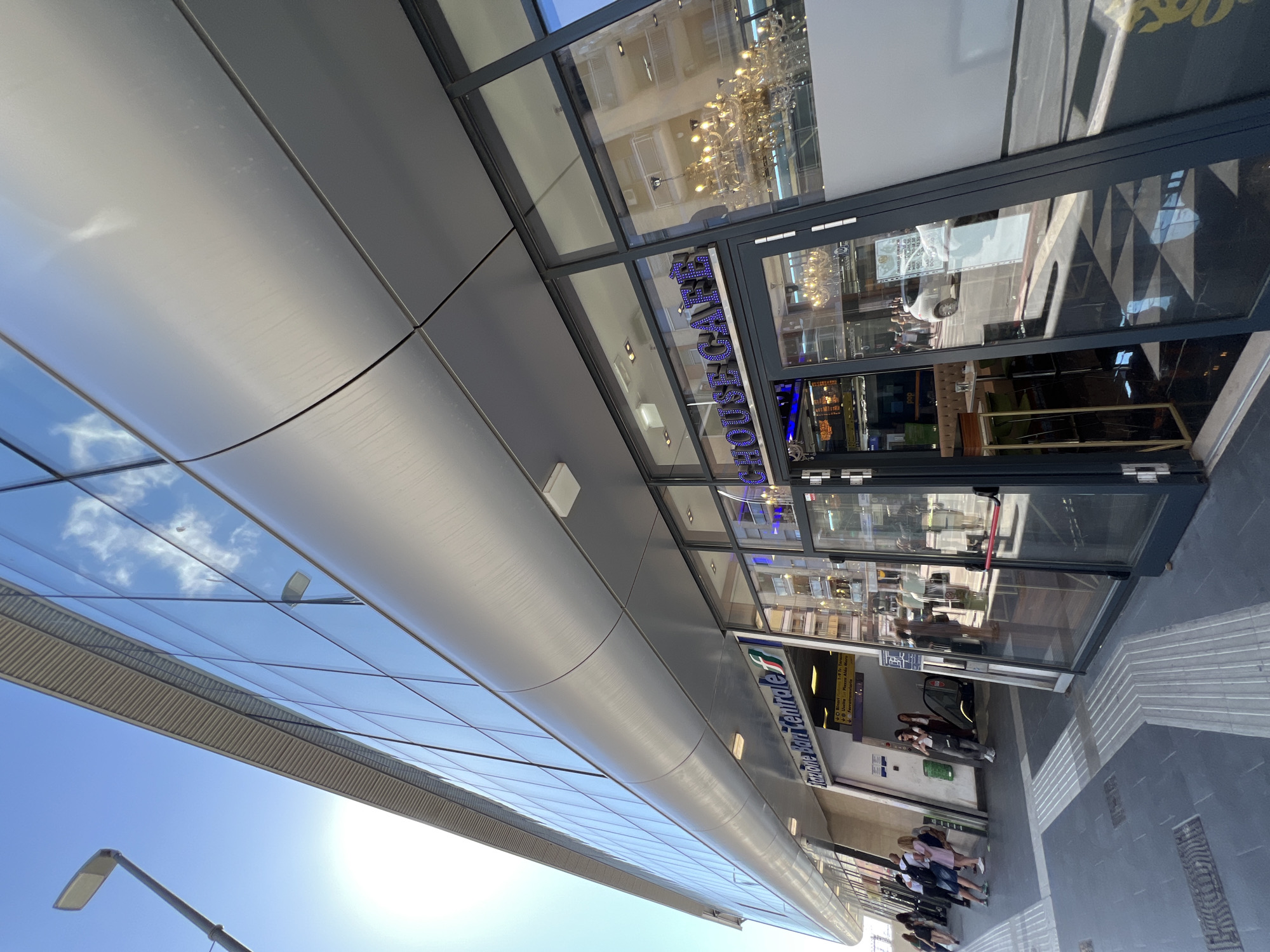 The image captures a vibrant, sunny street scene featuring the modern facade and entrance of a building, likely a train or bus station, given the prominent signage. The viewpoint is from a low angle, looking upwards towards the building's sleek, curved metallic panels and extensive glass windows.
Several people are visible near the entrance, appearing to enter or exit the building. They are casually dressed and engaged in typical public transit activities. On the paved sidewalk leading to the entrance, tactile paving for the visually impaired is present.
A key detail identifying the location is the visible text "Bari Centrale" displayed on a sign above the entrance, accompanied by what appears to be the Trenitalia (Italian national railway) logo. This confirms the setting as Bari Centrale, Italy. Above the main entrance, reflected in the glass, a blue neon sign reads "HOUSE CAFE," suggesting a food or beverage establishment within.
The weather is clear and sunny, indicated by the bright sky and the sun's glare in the lower left portion of the image. Reflections of clouds and blue sky are visible in the building's glass panels, creating interesting visual patterns. Through the glass, glimpses of the interior reveal an escalator or stairs, and reflections of an older building across the street can also be seen. Interior lighting, including chandeliers, is visible through the windows of the "HOUSE CAFE" area. The overall impression is one of a busy, modern urban transport hub during the daytime.