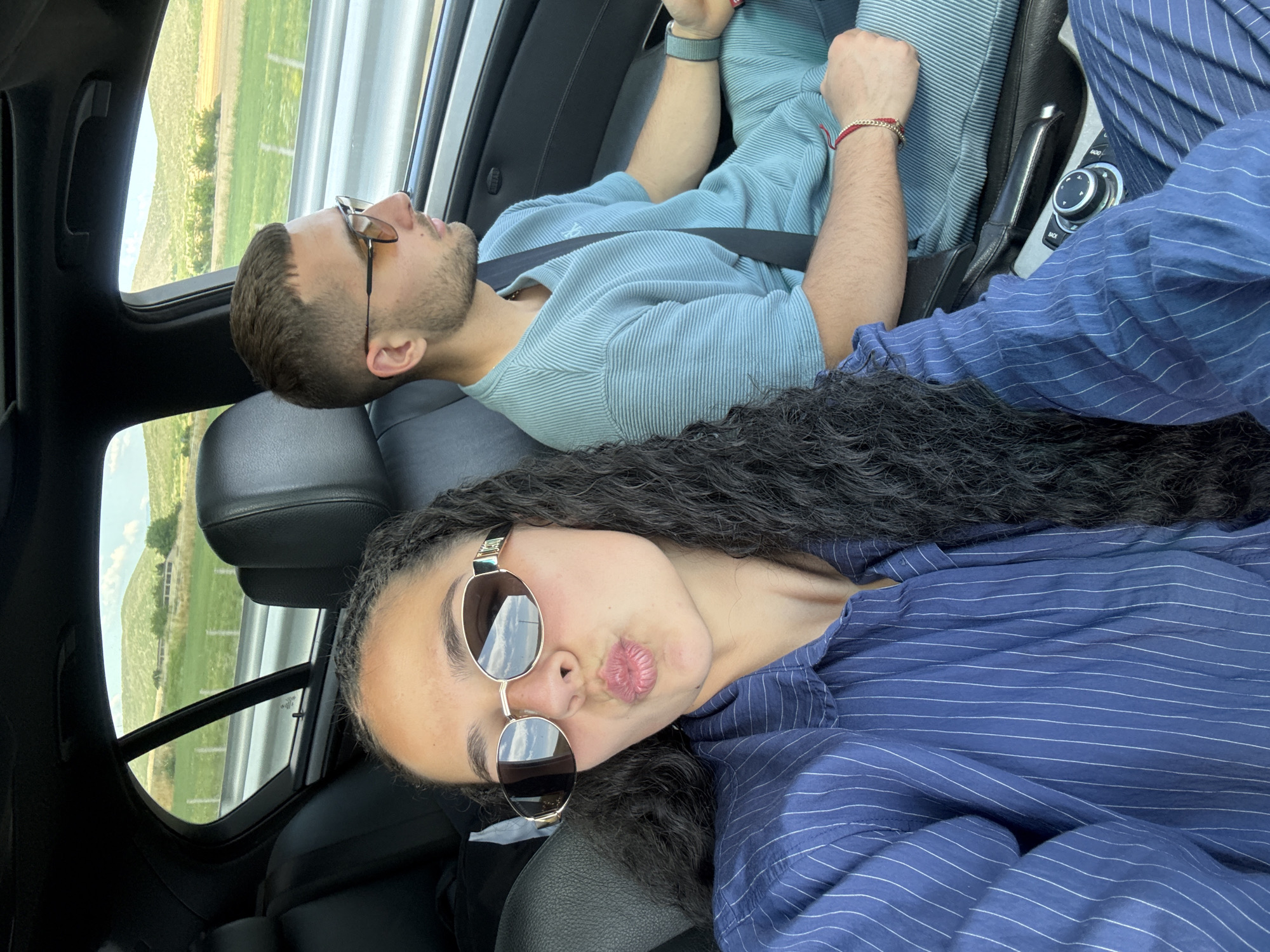 A medium shot from the interior of a car shows two people. In the foreground, a woman with long, dark curly hair and wearing a blue pinstripe shirt and oval sunglasses puckers her lips towards the camera. To her right, a man with short brown hair, a beard, and sunglasses wears a textured blue t-shirt. He is looking to his right. The car's interior includes black leather seats and a center console with a dial. Through the car window, a rural landscape with green hills and a cloudy sky is visible. The location is Polsko Padarevo, Bulgaria.
