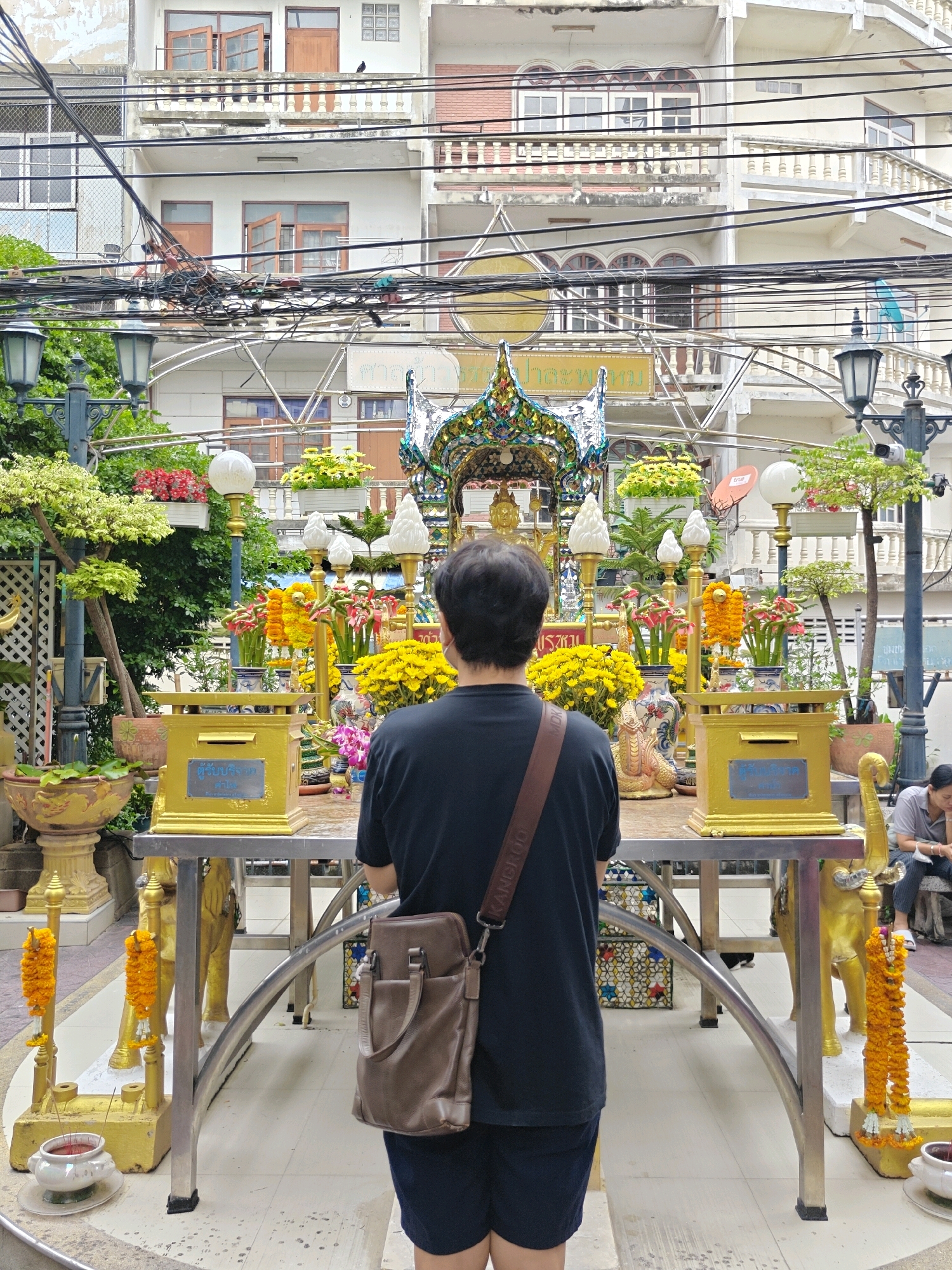 An outdoor scene in Bangkok, Thailand, features a man facing away from the camera, observing a shrine adorned with offerings of flowers and ornaments. The shrine is situated on a table with golden decorative elements, including statues of elephants. In the background, a multi-story building with balconies and numerous electrical wires is visible. To the right, a woman sits on a bench, looking down. The ground is paved with tiles, and potted plants and decorative garlands are placed around the shrine.