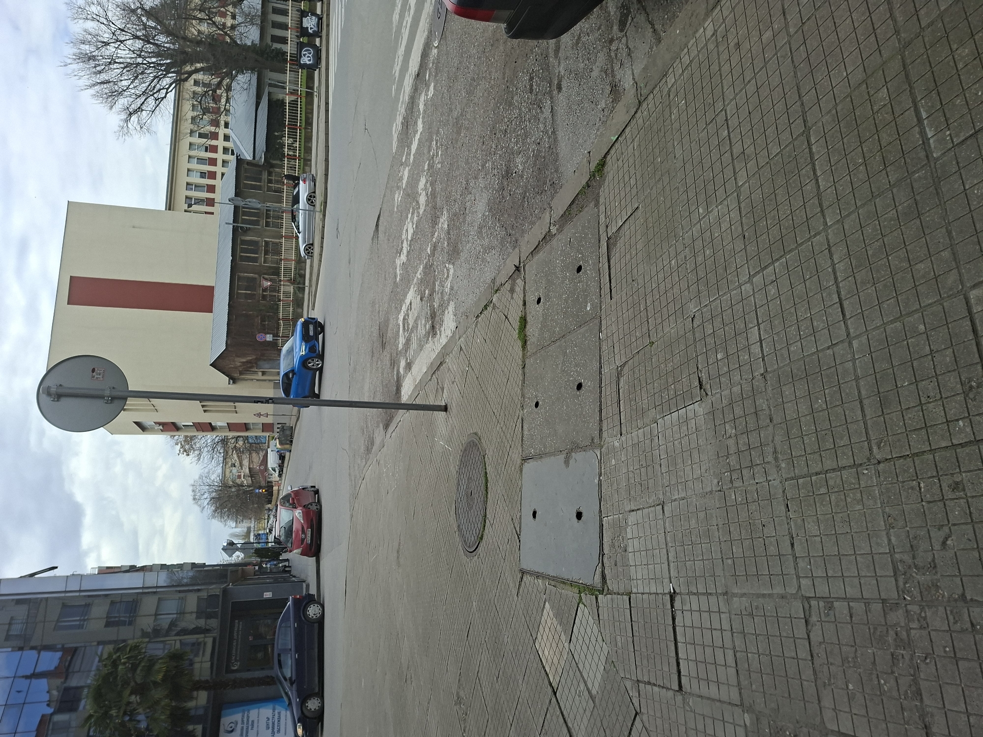 The image shows an urban scene in Plovdiv, Bulgaria. The perspective is ground-level, tilted slightly upward. The foreground is dominated by a tiled sidewalk made of square gray stones. A portion of the road is visible, with a crosswalk painted on it. Several cars of various colors (blue, red, dark gray) are parked or moving along the road. A traffic sign is present on the left side of the image. Buildings line both sides of the street, exhibiting diverse architectural styles from older constructions to modern glass-fronted buildings. Bare trees are visible in the background. The sky is overcast. A manhole cover is visible on the sidewalk.