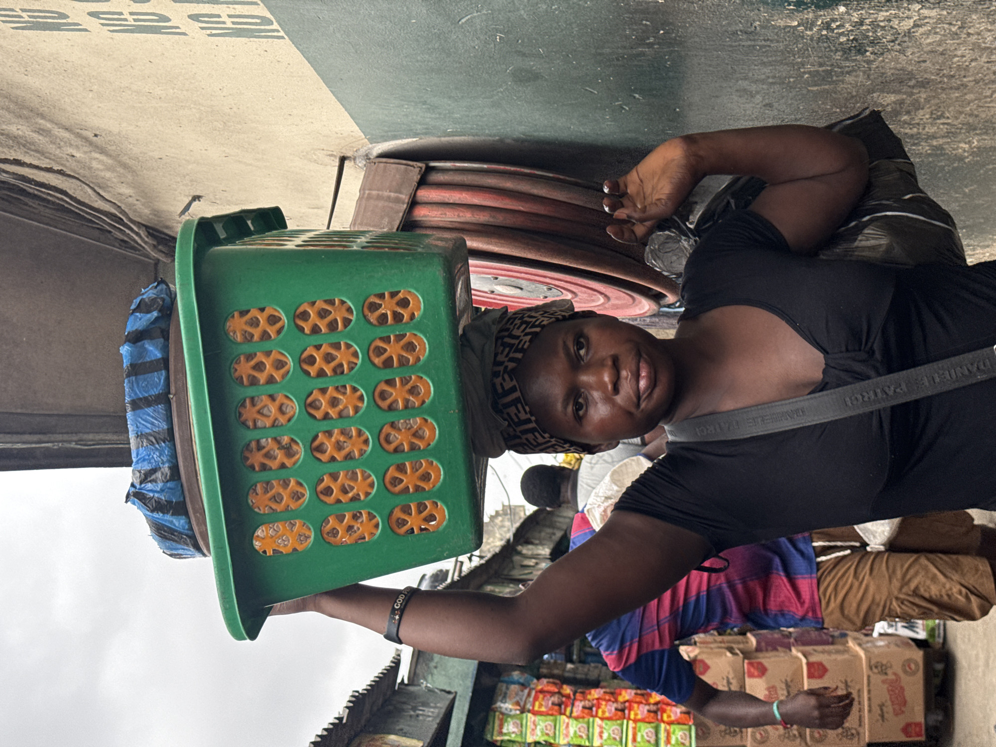 In a busy outdoor market in Lagos, Nigeria, a woman balances a large green plastic basket filled with goods on her head. The basket, which has orange-colored openings, contains items wrapped in blue and black plastic. She wears a black t-shirt, a patterned headwrap, and a bracelet on her left wrist. Behind her, a man in a blue and red striped shirt and brown shorts is visible, along with stacks of boxed goods. The environment appears to be a street with a textured, light-colored wall to the right, adorned with vertical green lettering. A coiled red hose is mounted on the wall. The sky is overcast.