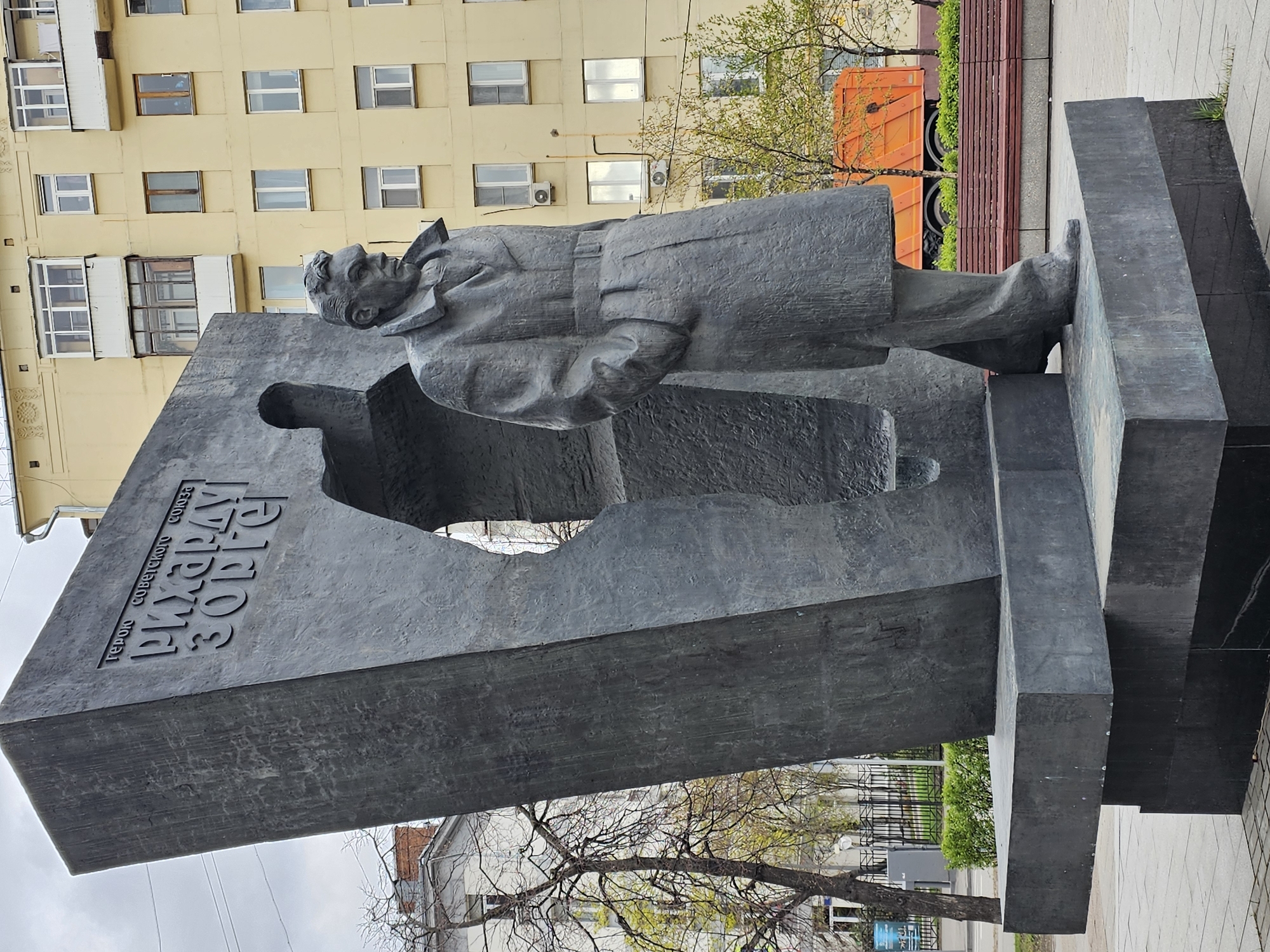 This image captures a monument dedicated to Richard Sorge, located in Moscow, Russia. The central subject is a large, dark-colored statue of a man, depicted in a long coat with a serious, upward-gazing expression. The statue stands before a substantial, dark grey slab of material, which features a cutout shape behind the figure, possibly representing another person or a symbolic void.
On this large slab, Cyrillic text is prominently inscribed: "ГЕРОЮ СОВЕТСКОГО СОЮЗА РИХАРДУ ЗОРГЕ," which translates to "TO THE HERO OF THE SOVIET UNION RICHARD SORGE." The monument's base is made of dark, polished stone blocks, resting on a paved area, suggesting a public square or park.
In the background, a light yellow multi-story apartment building with numerous rectangular windows and some enclosed balconies is visible. Its architecture suggests a typical Soviet-era design. Trees with a mix of bare branches and burgeoning green leaves frame the scene, indicating the season is likely spring. Partially visible behind the monument's base and trees is a bright orange utility truck and elements of urban landscaping like wooden benches and fencing. The sky appears overcast, providing soft, diffused lighting, typical of daytime. The monument's rugged texture and imposing scale convey a sense of solemnity and historical importance.