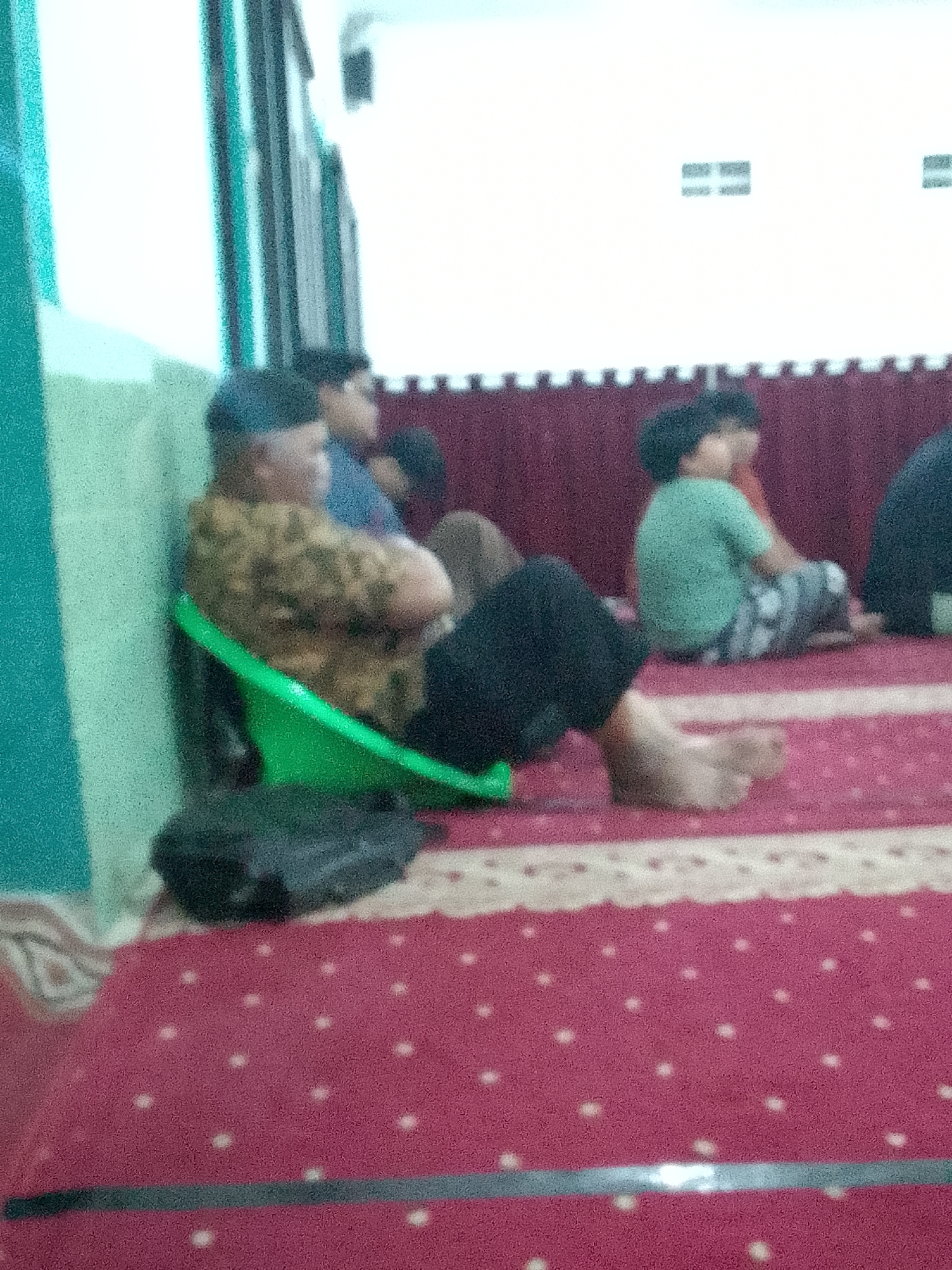 In Gunungmasigit, Indonesia, a group of individuals are gathered indoors. A man wearing a patterned brown shirt and a black cap sits in a green plastic chair. His arms are crossed, and his bare feet are visible. Beside him, a dark bag rests on the red carpeted floor. In the background, several children and adults are seated on the floor, facing away from the camera. One child wears a light green shirt and patterned pants. The room is brightly lit, with a white wall and what appear to be decorative elements on the wall. A burgundy curtain hangs in the background. The image quality is blurry, suggesting it may have been taken with a low-resolution camera or under low light conditions.