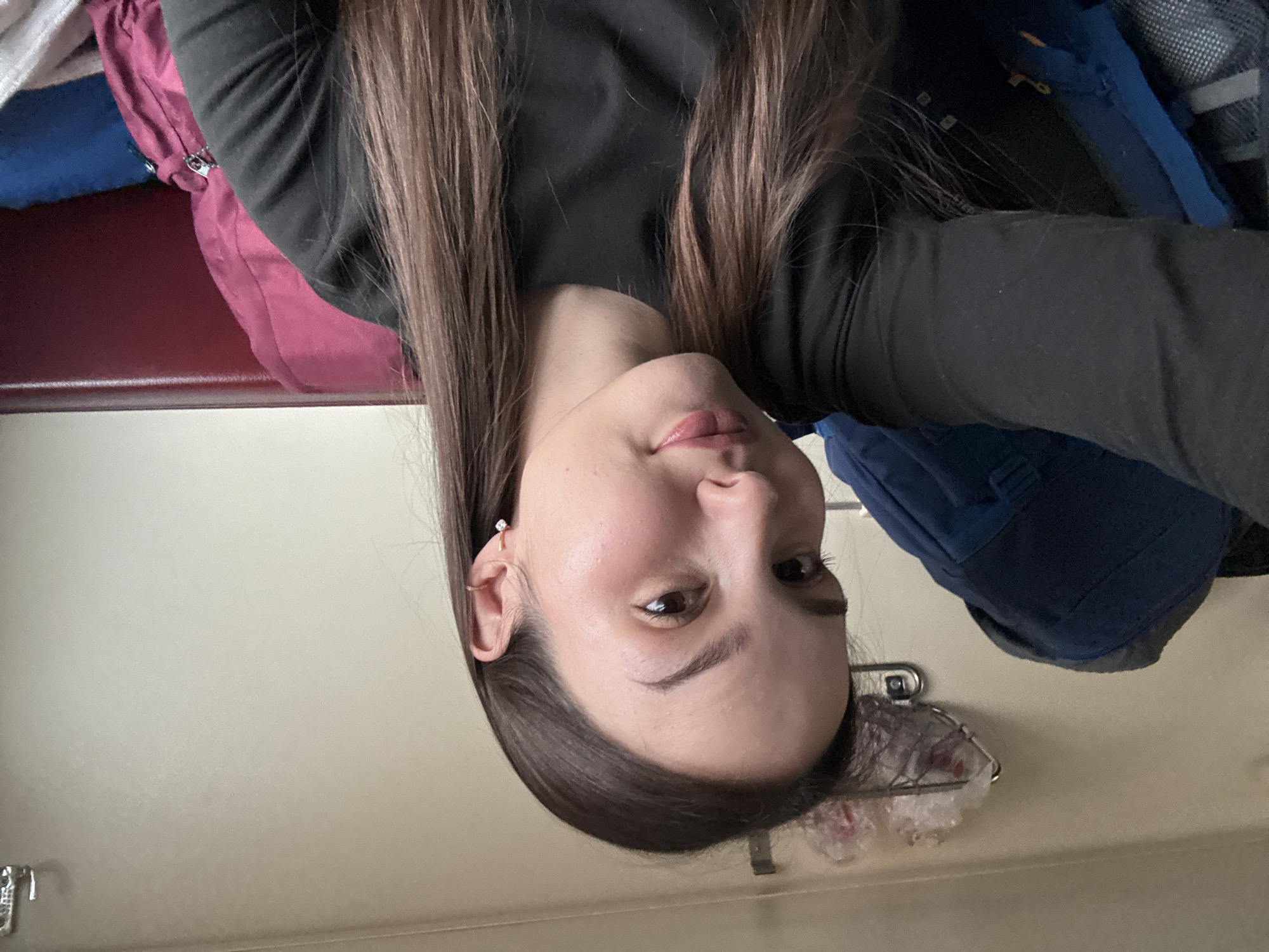 The image captures a selfie of a female individual inside what is consistent with a Russian train compartment, with the contextual location identified as Usolye-Sibirskoye, Russia. The subject is a young adult female, positioned centrally, looking directly into the camera with a neutral expression and a slight upward curve to her lips. She has long, straight, dark brown hair with lighter ends, parted down the middle, extending over her shoulders. She wears a dark, long-sleeved top. A small, clear-stone stud earring is visible in her right earlobe, along with a small ring in her right helix piercing.
To the subject's left, a large, dark blue backpack is visible. Above and behind her left shoulder, a metal wire rack is affixed to a light-colored wall, containing multiple translucent plastic bags. A silver metal hook is mounted higher on the wall to the right. To the subject's right, a padded maroon surface, consistent with a train seat or headrest, forms the background. Against this, a fuchsia-colored soft bag with a visible silver zipper is partially obscured by the subject. Below the fuchsia bag, a blue object and a white fabric item are partially visible. The immediate environment suggests a confined travel space within a passenger train car.