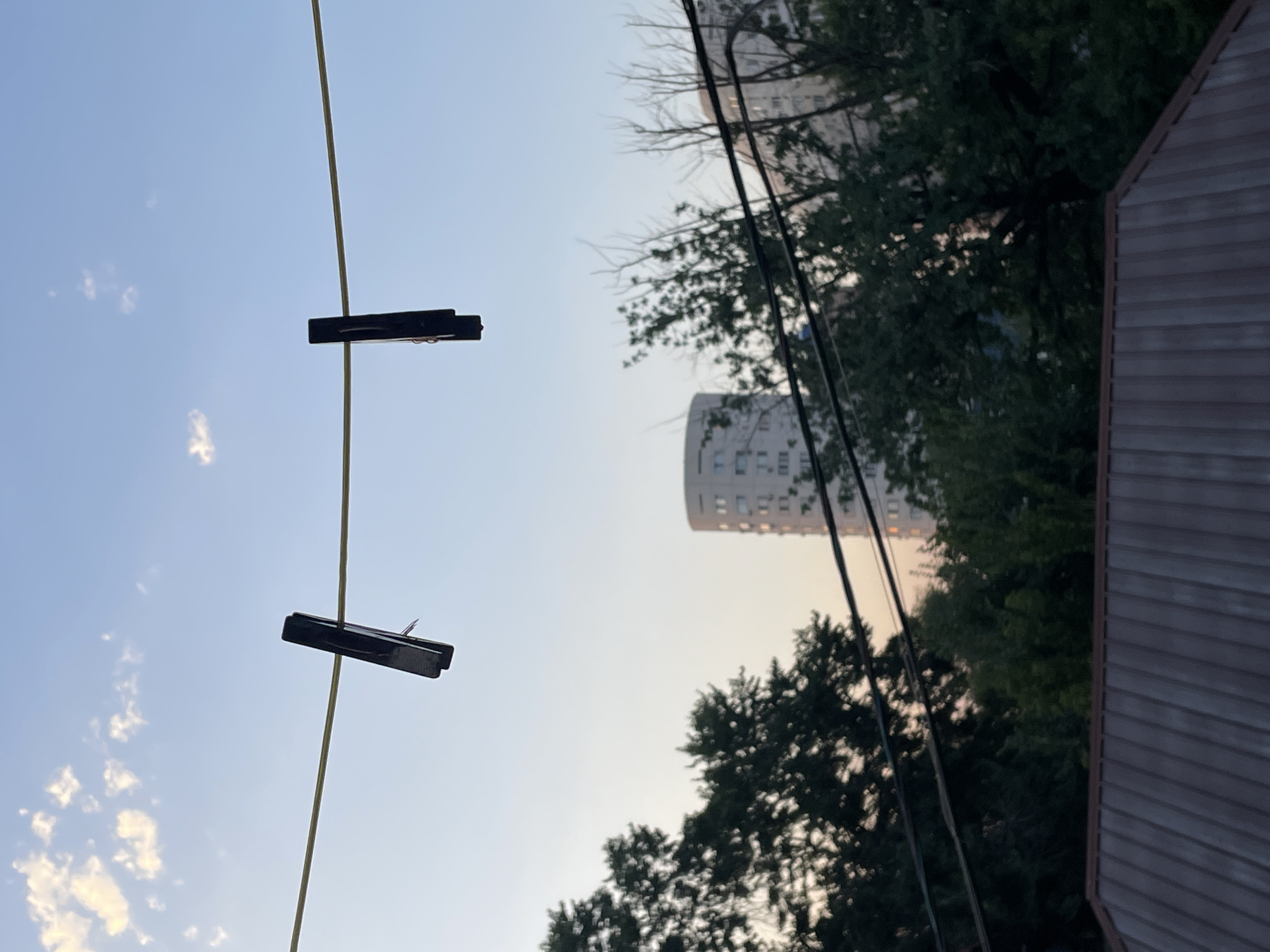 Two black clothespins are attached to a clothesline, which is strung horizontally across the frame against a clear blue sky with scattered white clouds. In the middle ground, a cylindrical building with numerous windows is visible through a canopy of green trees. The trees are dense and dark, obscuring much of the background. Below the trees, a dark brown, corrugated metal roof takes up the bottom portion of the image. The lighting suggests it is either early morning or late afternoon, with soft, diffused light. The image appears to be taken from a residential area, possibly a balcony or backyard, with a city skyline in the distance. There are no people visible in the image. The overall impression is one of domestic life set against an urban backdrop. The image is taken in Almaty, Kazakhstan.