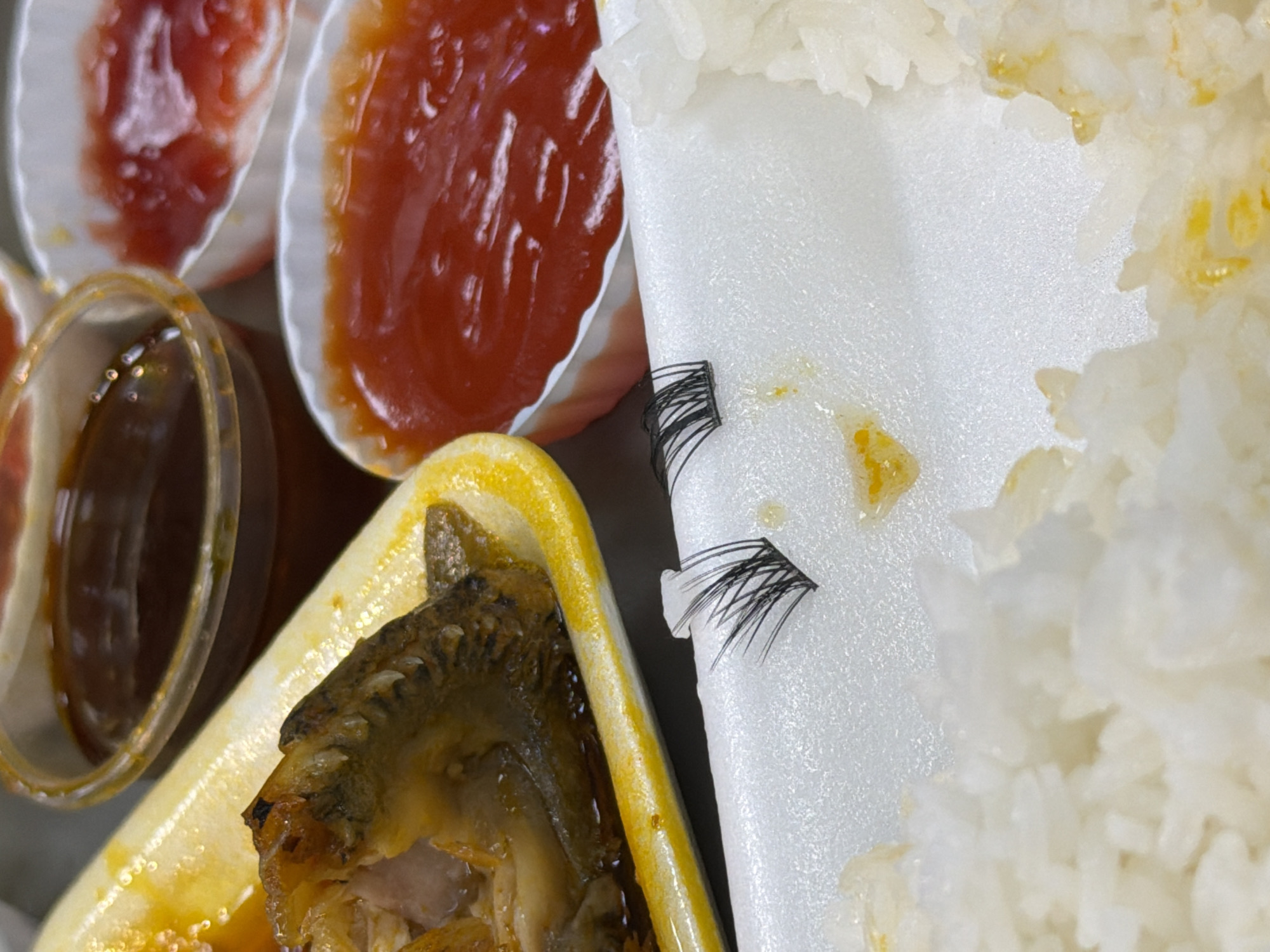 A close-up shot shows a food container with rice, fish, sauces, and a pair of false eyelashes on the styrofoam divider. The food appears to be a takeout or delivery meal. The rice is clustered on the right side of the divider. The fish, in a yellow tray, is positioned in the lower left of the frame. Small containers of red and brown sauces are visible at the top left. The false eyelashes are affixed to the styrofoam surface. No people are visible in the image. The location in Đà Nẵng, Vietnam, is not determined.