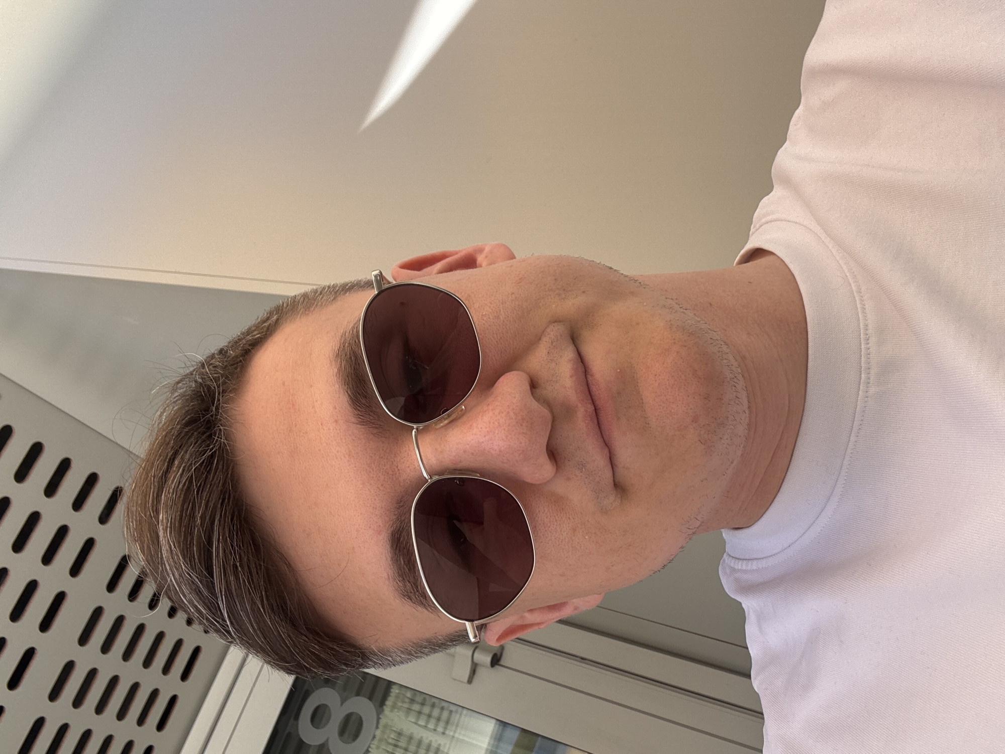 A close-up, slightly angled photograph shows a person with light skin, wearing sunglasses and a white t-shirt. The person has short hair, with some gray strands visible. The sunglasses have a thin silver frame and dark lenses. The background appears to be an indoor or semi-outdoor space, with a wall featuring a pattern of vertical slots, possibly part of a ventilation system or architectural design. Part of a window and other indistinct elements are visible through the sunglasses' lenses. The location is not specified.