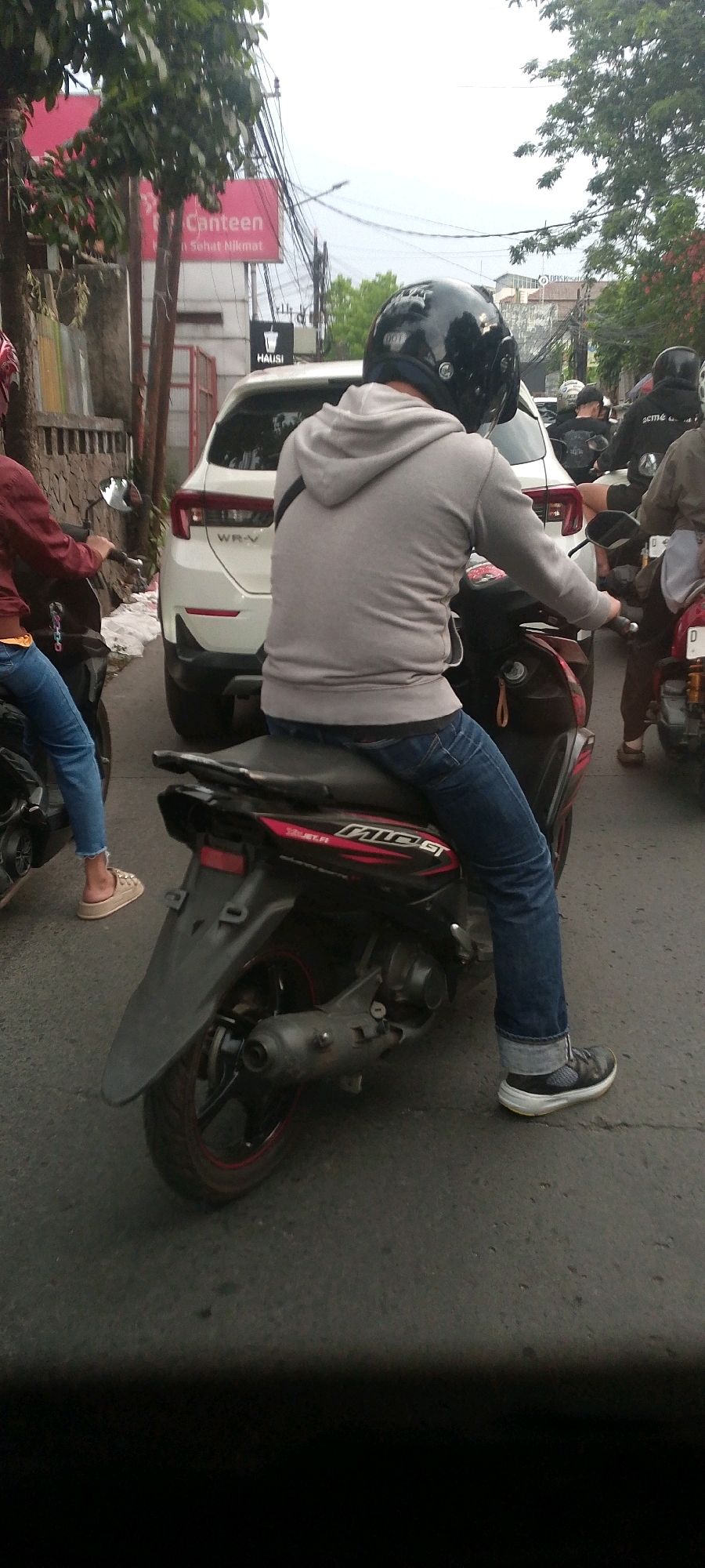 The image shows a street scene in Bandung City, Indonesia, with several people on motorcycles in traffic. In the foreground, a person wearing a gray hoodie and blue jeans is riding a dark-colored scooter. The scooter has red and black accents. Behind this scooter, a white Honda WR-V car is visible. Further in the background, other motorcyclists are present, including one wearing a black jacket and another in a light brown jacket. Overhead, a complex network of electrical wires is strung across poles. A red sign with white text reading "Canteen" and "Sehat Nikmat" is visible on a building to the left, along with a black sign for "HAUSI." The overall environment appears to be a busy urban street during daylight hours.