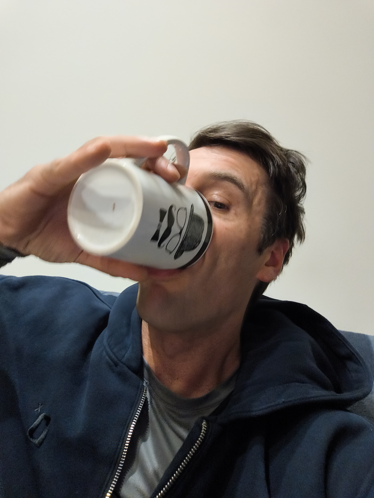 A man is drinking from a white mug with a black design of a hat and glasses on it. He is wearing a blue zip-up hoodie over a gray t-shirt. The background is a plain white wall. No information indicates this is Moscow, Russia.