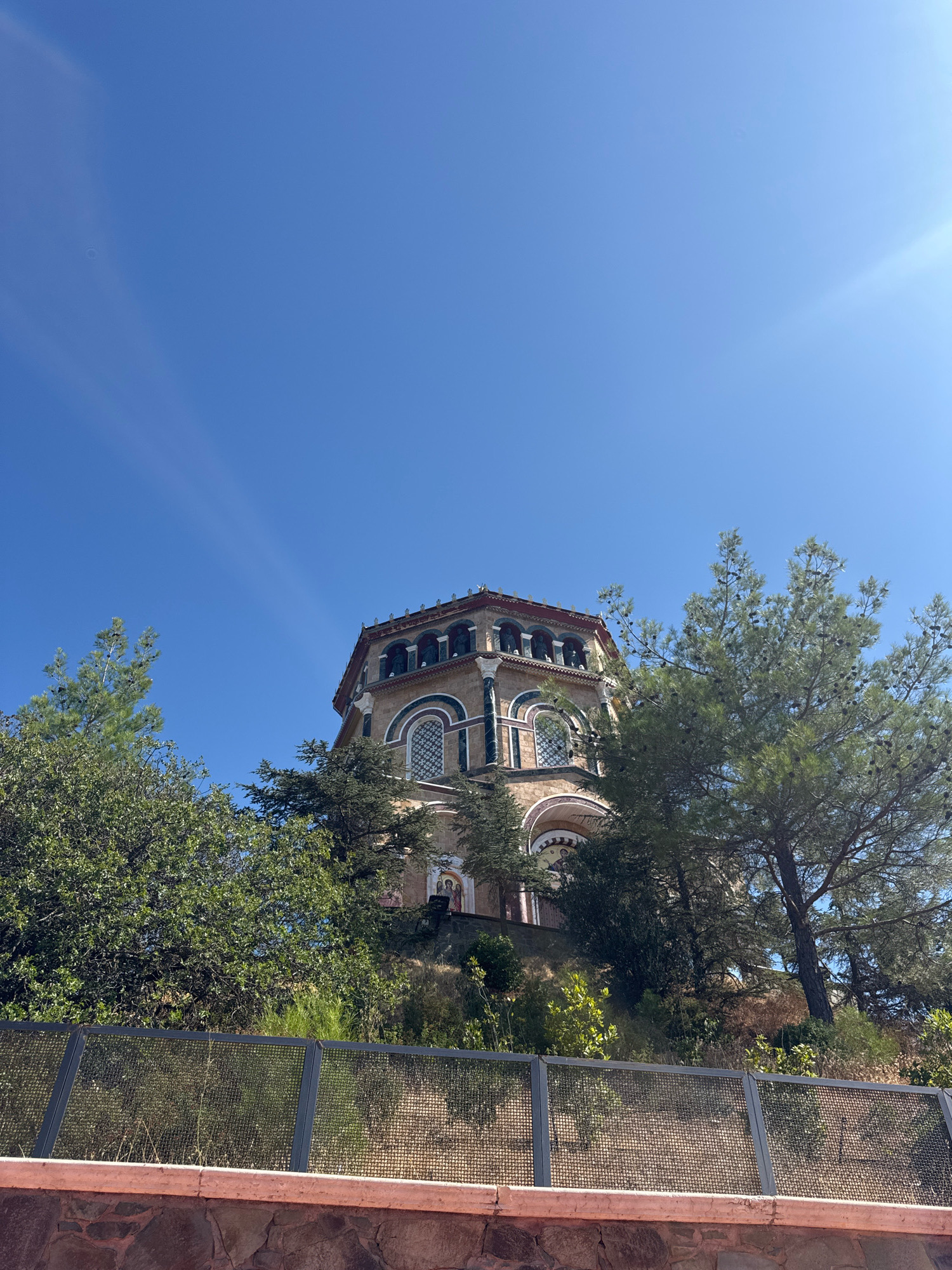 The image shows an exterior view of the Church of the Holy Cross in Gerakies, Cyprus, on a clear, sunny day. The church, constructed from stone, is situated on a hill and features a distinctive octagonal shape with a red-tiled roof and arched windows. It is partially obscured by lush green trees, including pine trees with visible cones. In the foreground, there is a stone wall topped by a metal fence. The sky is a vibrant blue with subtle sunbeams visible. There are no people visible in the frame.