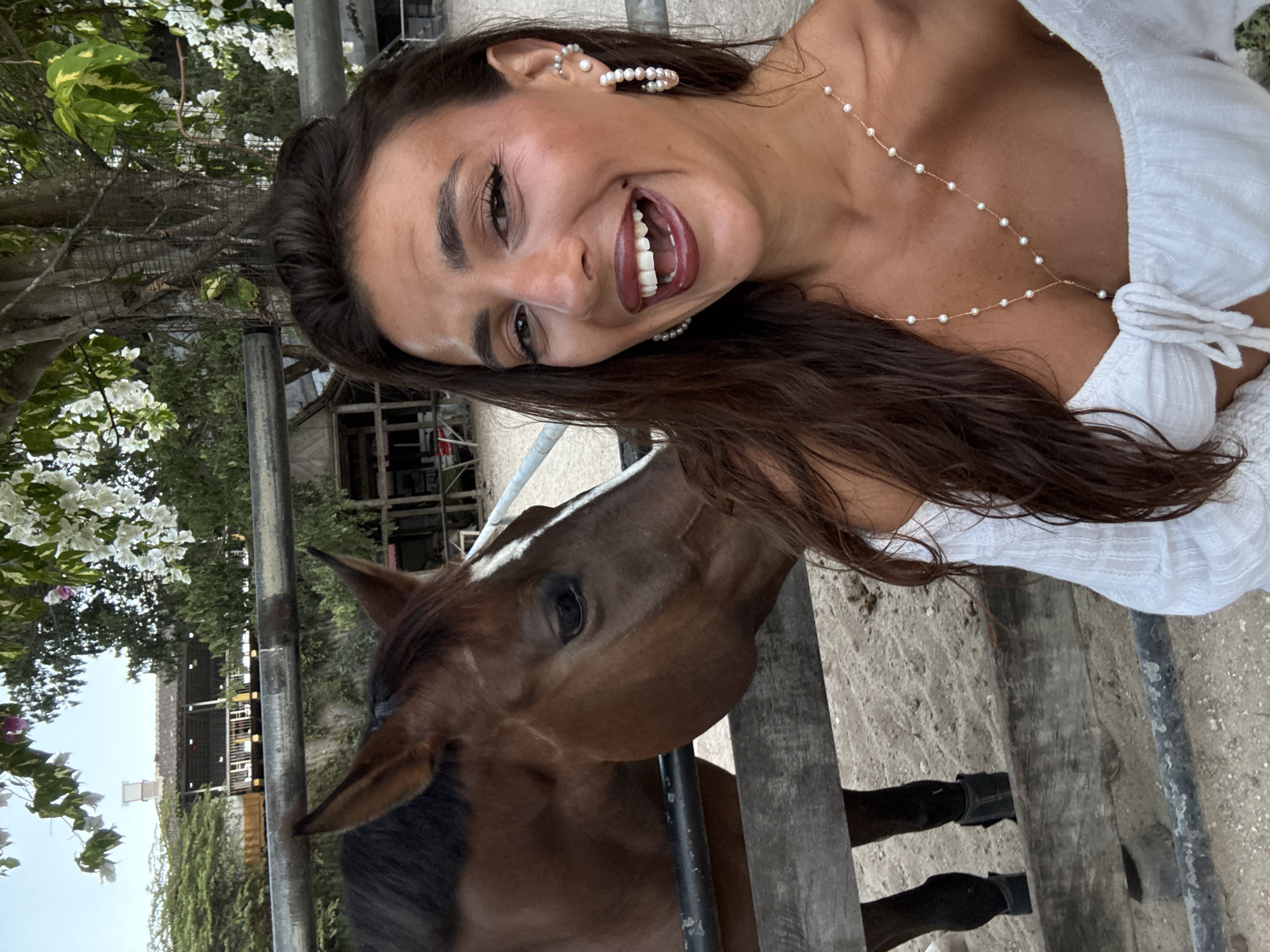 The image captures a selfie-style photograph of a woman and a horse in an outdoor setting, consistent with a stable or animal encounter area in Kuta Selatan, Indonesia. In the foreground on the right, a woman with long, dark brown hair and a tanned complexion smiles broadly towards the camera, her mouth open to reveal white teeth. She wears dark eyeliner, minimal eye makeup, and dark red lipstick. Her attire consists of a white, off-the-shoulder, textured top with a tied front. Accessory details include multiple pearl-themed pieces: hoop earrings with pearls, a pearl cuff earring on her upper left ear, and a delicate gold-colored necklace with small pearls spaced along its length, forming a Y-shape descending onto her décolletage.
In the mid-ground on the left, behind a dark metal upper rail and a wooden lower rail, stands a brown horse with a distinct white blaze on its face. The horse's head is turned towards the woman, its muzzle positioned near her shoulder, suggesting an interaction. Its dark eye is visible, and its ears are erect. The horse's lower legs, partially visible below the wooden fence, are dark and appear to have wraps or boots.
The background reveals a dirt or sandy ground within the enclosure. Beyond the fence line, lush green foliage and trees dominate, with cascades of white flowers (possibly bougainvillea) visible in the upper right and left corners. A multi-story building with a dark, tiled roof and a balcony or veranda supported by columns is discernible through the trees in the distance. The sky above is light blue, indicating daytime.