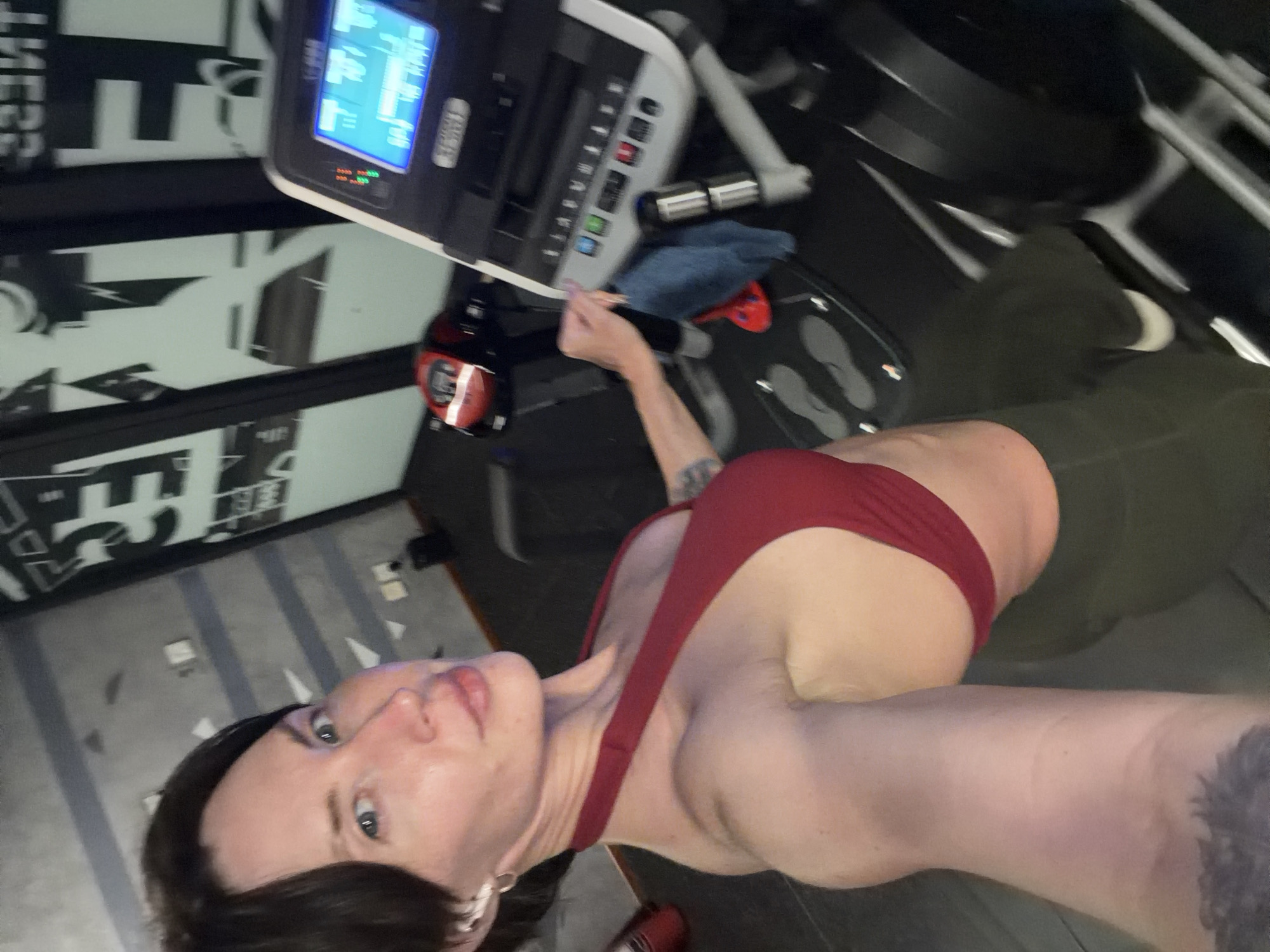 In a gym setting, a person with dark hair and fair skin takes a selfie. They are wearing a red halter-neck sports bra and dark green workout leggings. The person is standing in front of a treadmill, which has a digital display showing workout data. A blue towel is draped over the console of the treadmill. The background includes a wall with a large graphic design and a door or window. The overall lighting suggests an indoor environment, likely during daytime or with artificial lighting. The location is identified as Dong Ninh Hoa Ward, Vietnam, implying the gym is situated within this geographical area.