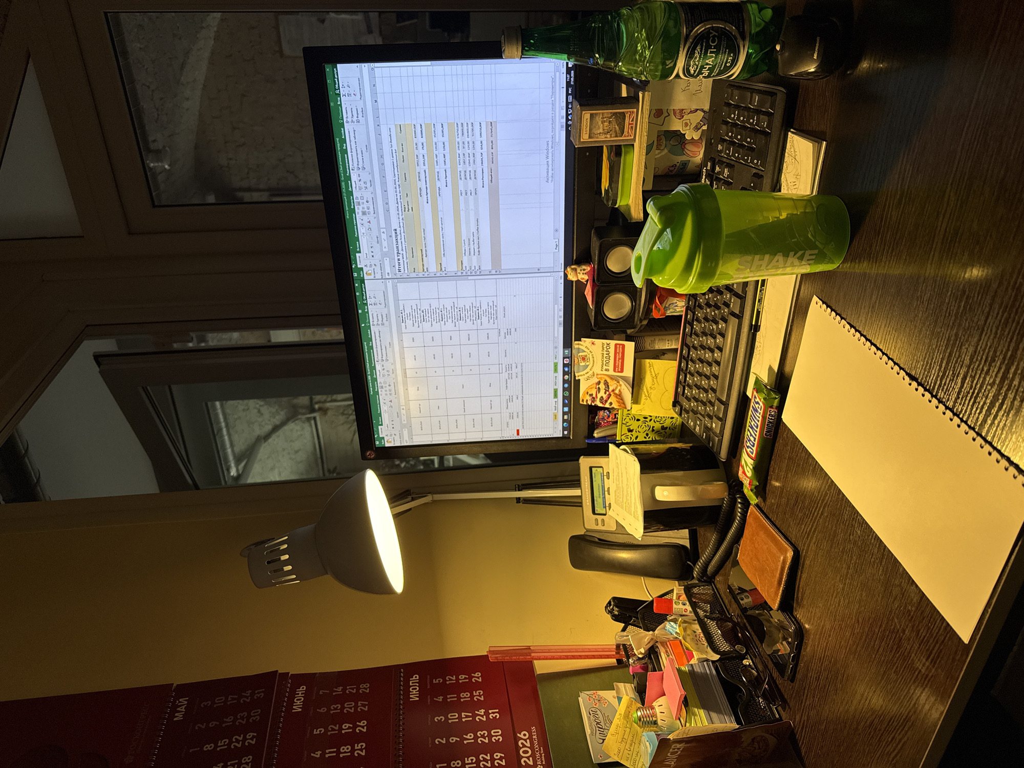 The image shows a desk setup in what appears to be a home office. A computer monitor displays an Excel spreadsheet, with a keyboard and a Microsoft mouse positioned below it. A green "SHAKE" protein shaker is placed to the right of the keyboard, near a notepad. Various items are scattered around the desk, including a bottle of "НАРЗАН" mineral water, speakers, small figurines, a box of sweets "В ПОДАРОК", a Snickers bar, a wallet, a pair of glasses, pens, sticky notes, and a desk organizer filled with stationery. A desk lamp is positioned to the left, illuminating the workspace. A calendar for May, June, and July 2026 is visible, with "ROSCONGRESS" printed on it. The calendar seems to be in Russian, which suggests that the location is in Russia. The presence of the "ROSCONGRESS" calendar likely hints that the image was taken in Saint Petersburg, Russia, as ROSCONGRESS is a major event organizer in that city. The window behind the monitor shows a glimpse of an exterior, hinting that the desk is placed next to a window. The overall lighting suggests the photo was taken in the evening or in a dimly lit room.