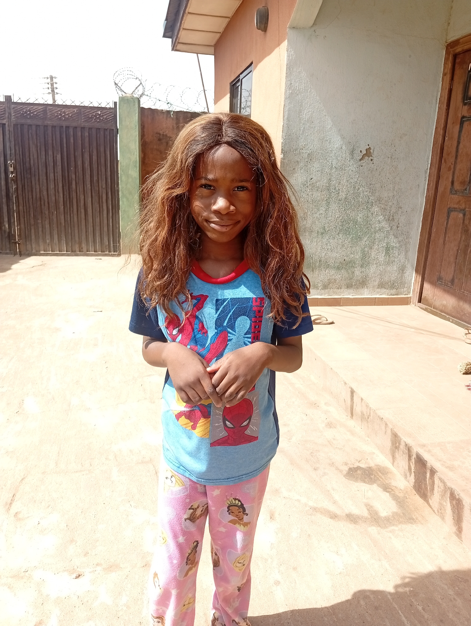 A young girl with brown hair stands in front of a building in Sabon Tasha, Nigeria. She is wearing a blue Spiderman t-shirt and pink princess-themed pajama pants. Her hands are clasped in front of her. Behind her is a concrete patio and the side of a building with a closed wooden door. To the left is a metal gate topped with barbed wire. The scene is bathed in bright sunlight.