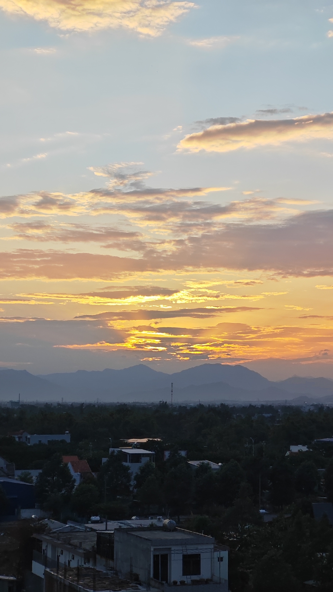 The image captures a panoramic view of a city at dusk, likely Đà Nẵng, Vietnam, characterized by its blend of urban development and natural scenery. The sky is dominated by a dramatic sunset, with vibrant hues of orange, yellow, and pink painting the clouds. Rays of sunlight pierce through the clouds, creating a striking visual effect against the backdrop of distant, hazy mountains. Below the sky, a dense canopy of green trees covers the middle ground, interspersed with residential buildings and some darker structures. In the foreground, the rooftops of several buildings are visible, showcasing various architectural styles. The overall impression is one of a serene evening cityscape, where the natural beauty of the sunset complements the urban landscape.